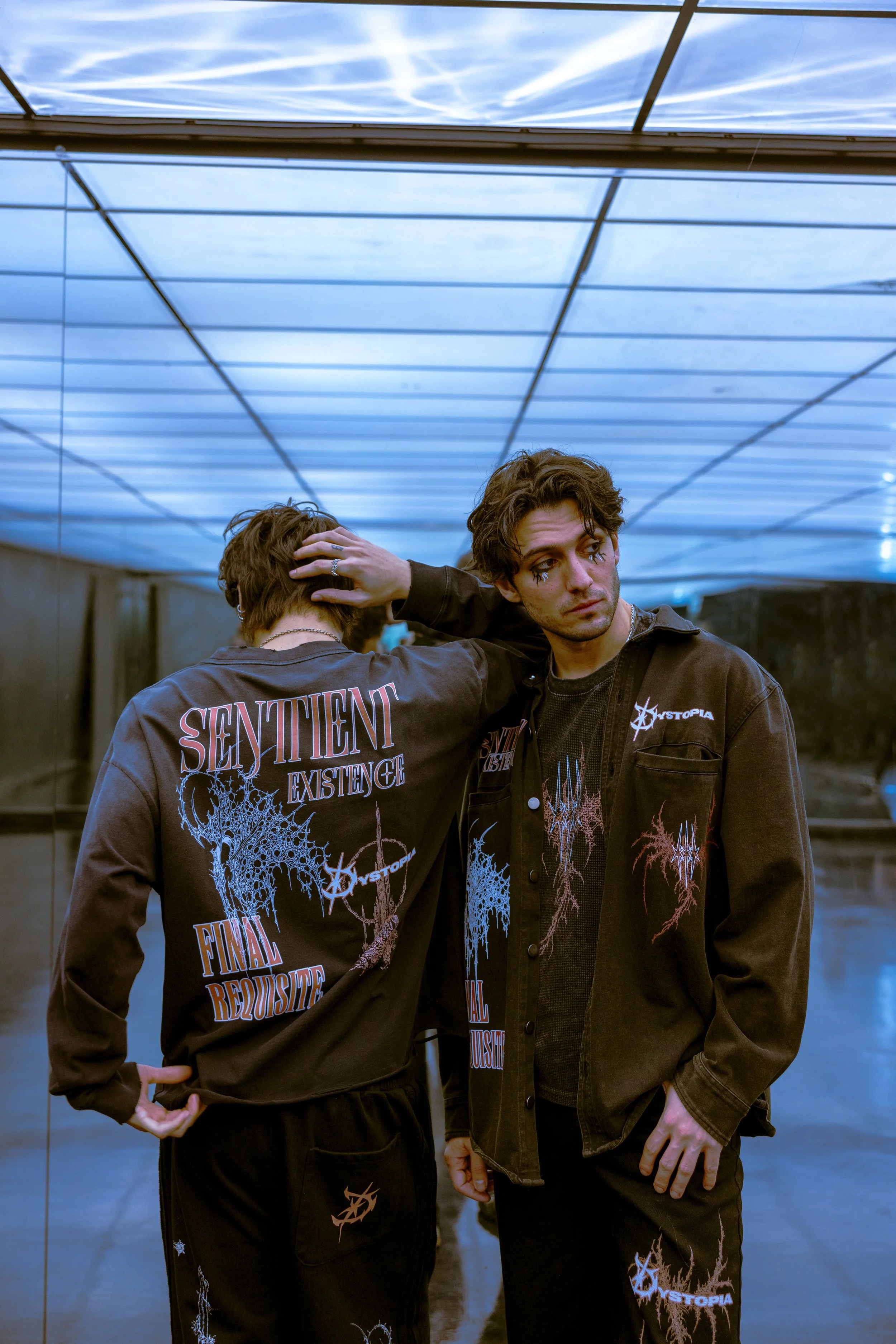 Two young men standing in a futuristic, reflective room with a blue-lit ceiling. They are wearing black jackets and pants with graphic designs and text related to the band Sentient and a theme of dystopia. One man faces the camera with a contemplativ