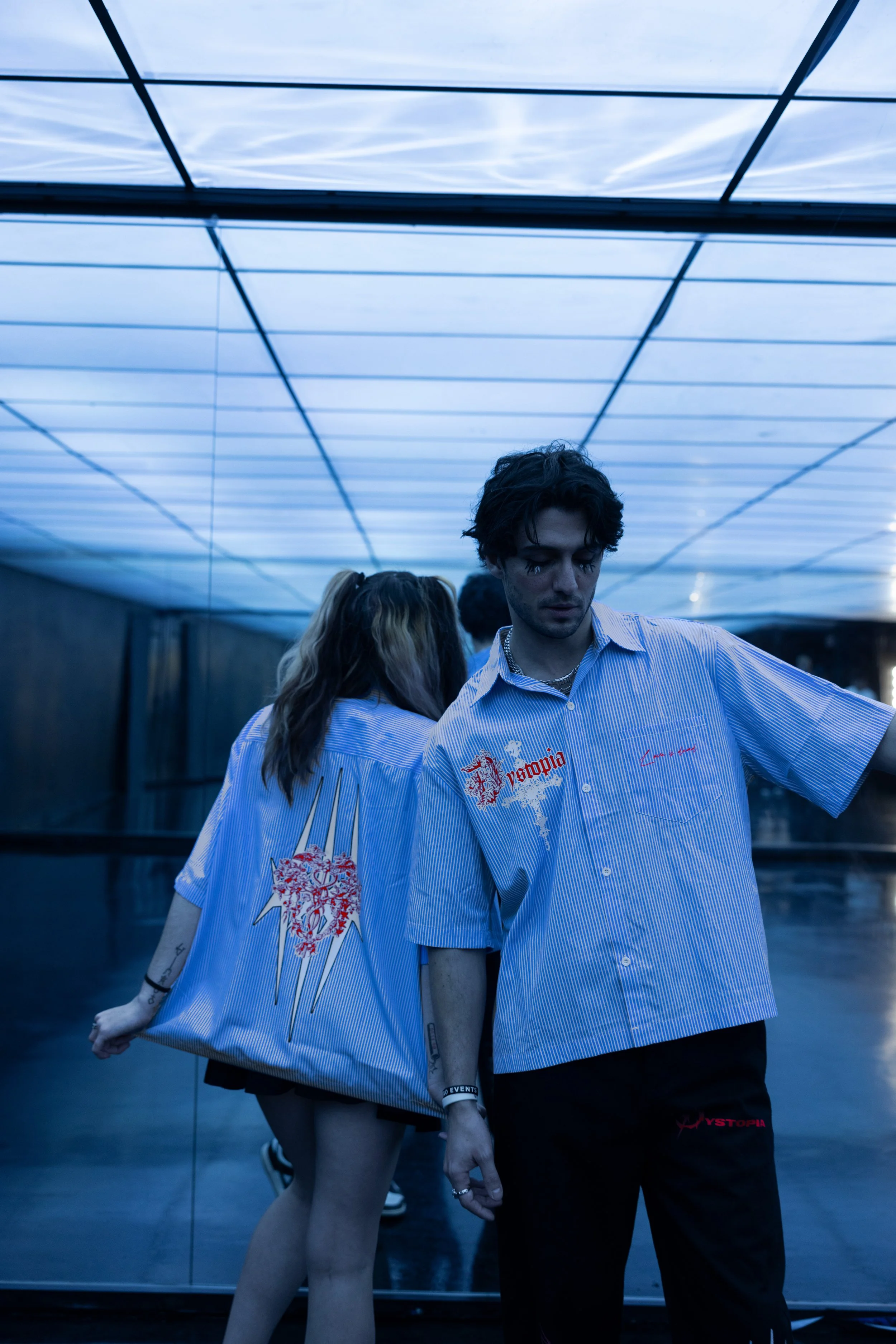 A young man and woman standing in front of a mirror, wearing matching striped shirts with graphic designs and text. The scene has a blue hue, and the background features a ceiling with reflective panels.