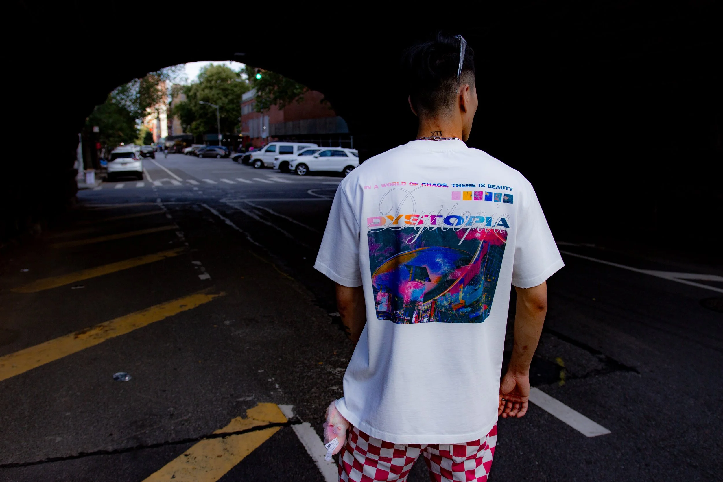 Person standing under a bridge or tunnel, facing away, wearing a white T-shirt with colorful graphics and text, and red and white checkered shorts, with a city street and parked cars visible in the background.