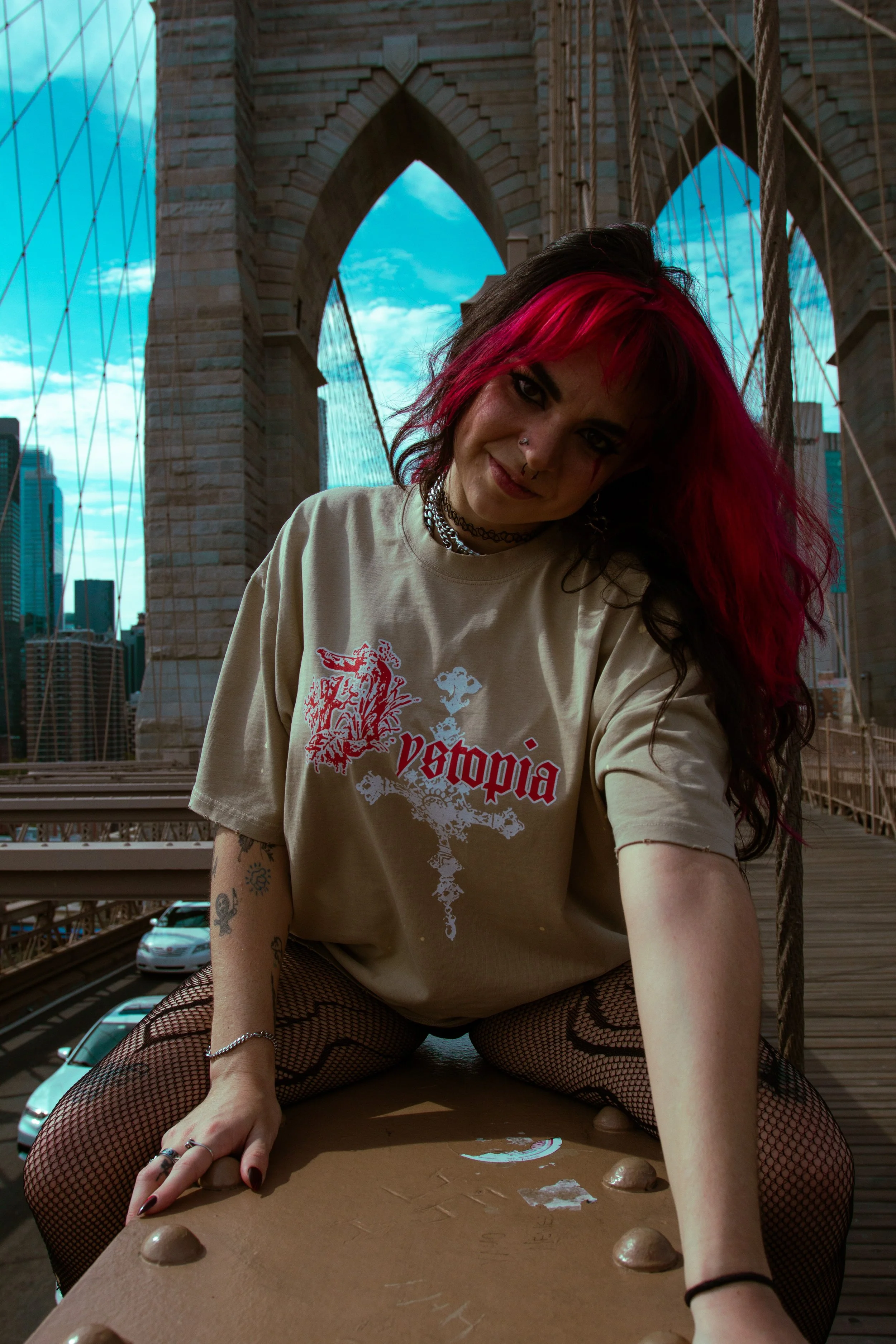 Young woman with pink and black hair sitting on a bridge, wearing a beige t-shirt with red and white text and graphics, fishnet stockings, and jewelry, with the Brooklyn Bridge and city skyline in the background.