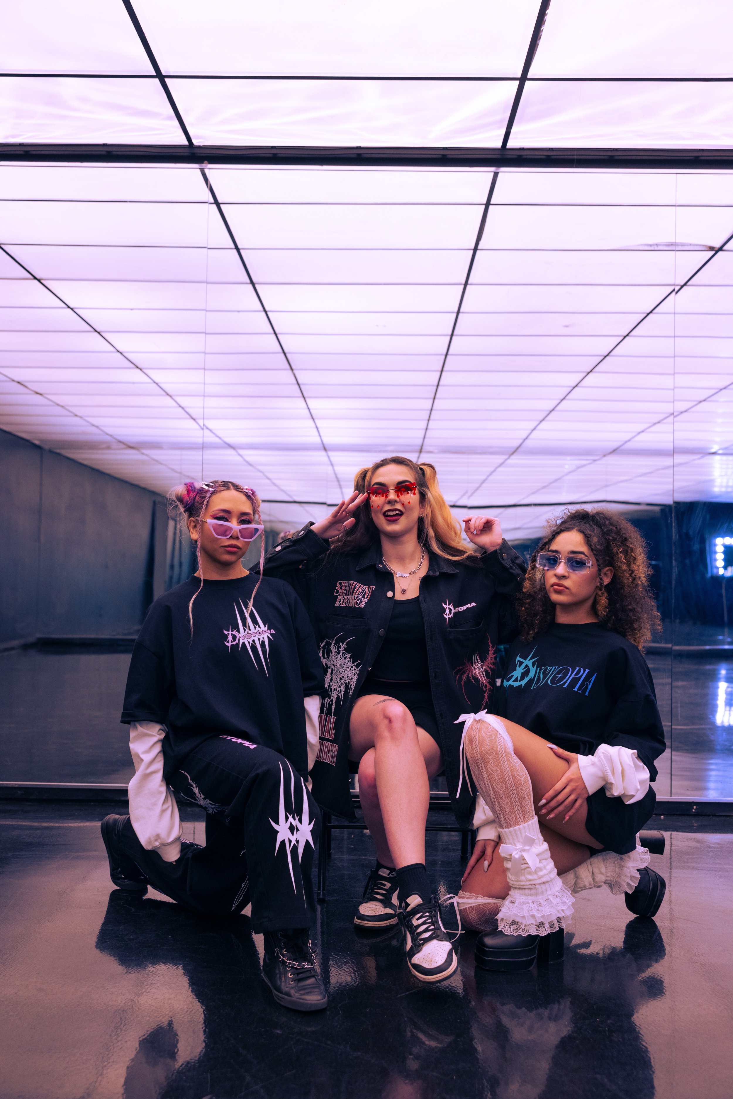 Three women in edgy, colorful outfits pose in front of a large mirror and purple-lit ceiling in a dance studio or nightclub.