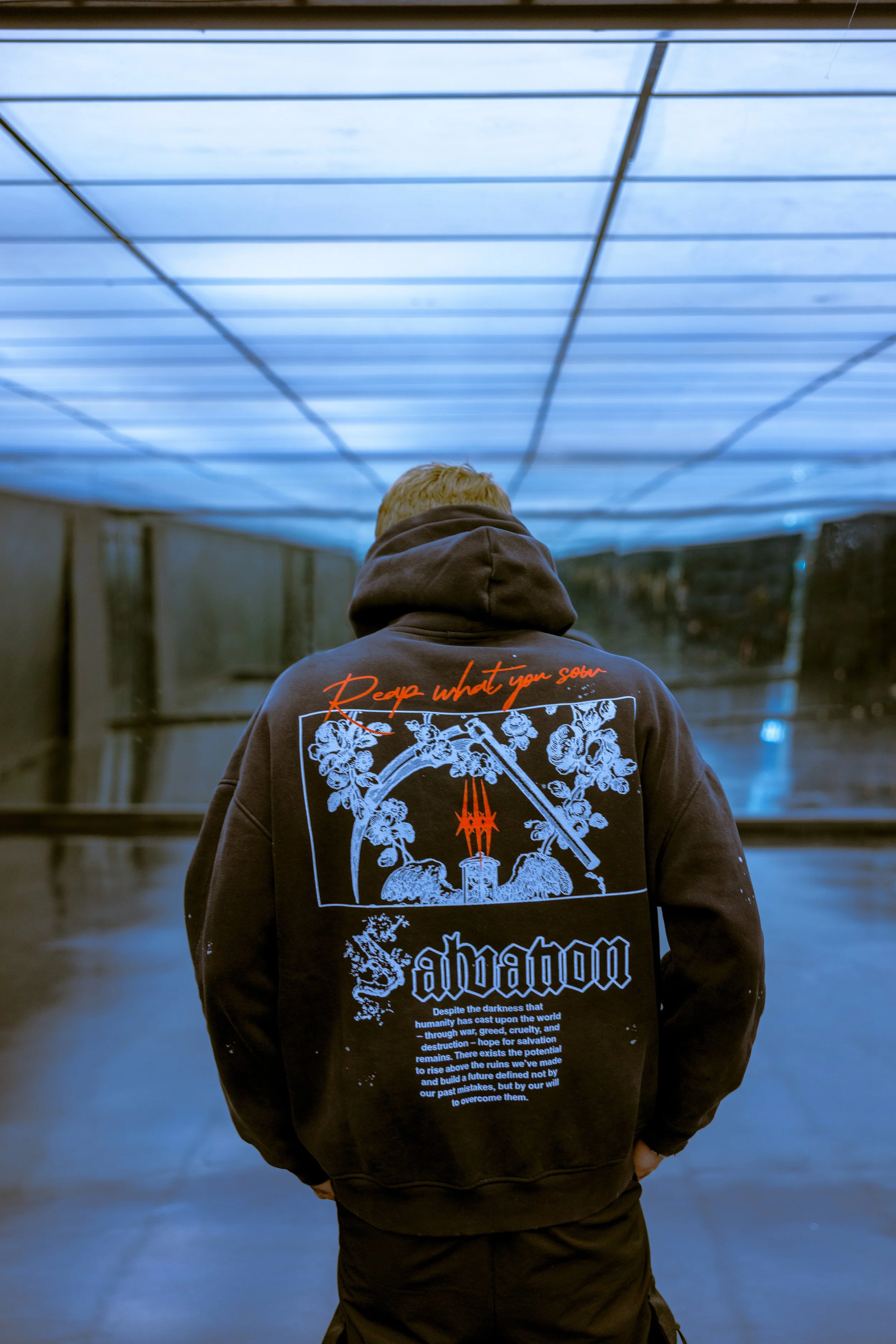 A person wearing a black hoodie with graphic designs, standing indoors under a blue-lit ceiling, facing away from the camera.