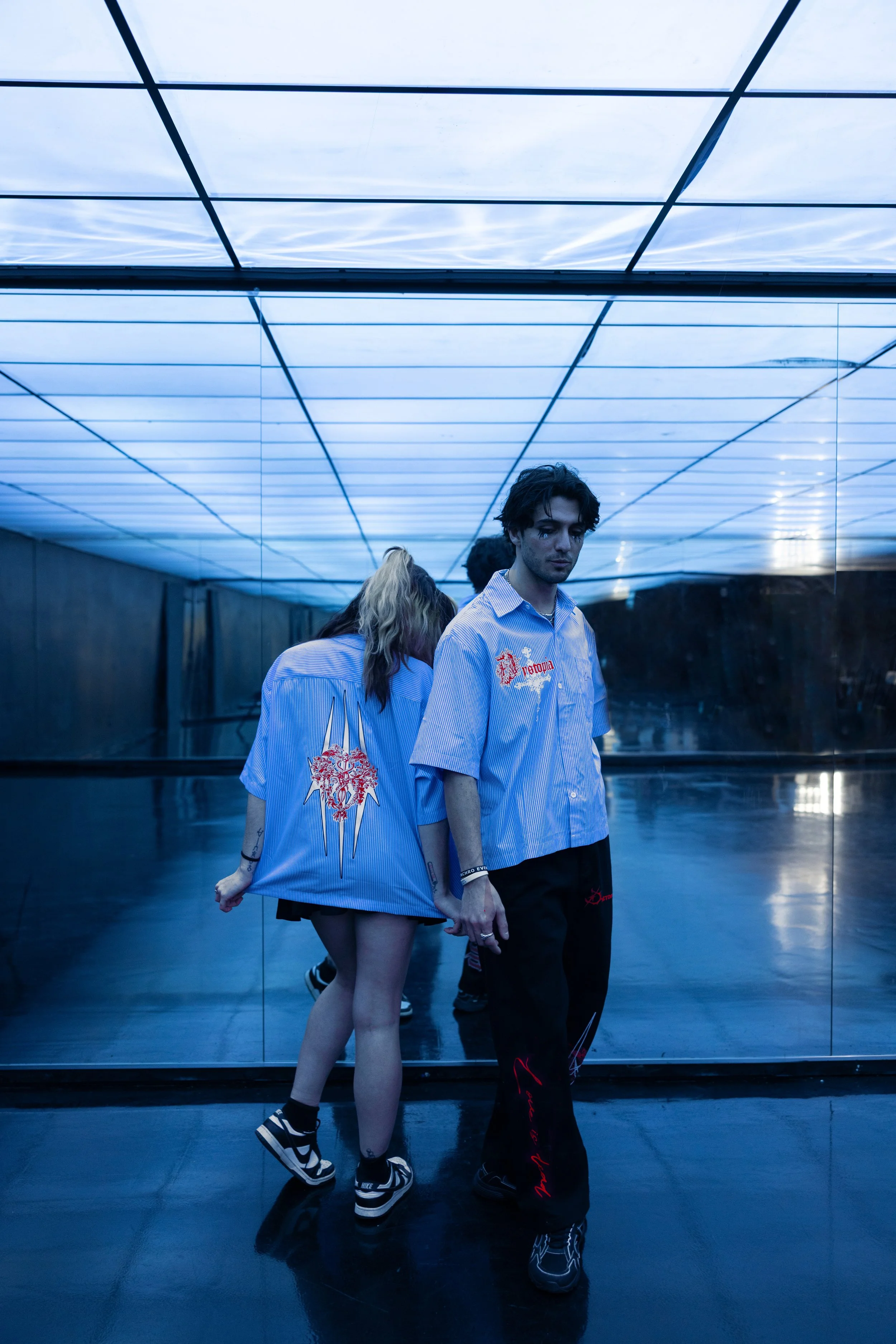 Two young people in blue shirts standing in front of a mirror in a dimly lit room with a blue ceiling and floor. The girl has long hair and is wearing shorts and sneakers, with her back to the mirror. The boy has dark hair and is facing the camera wi