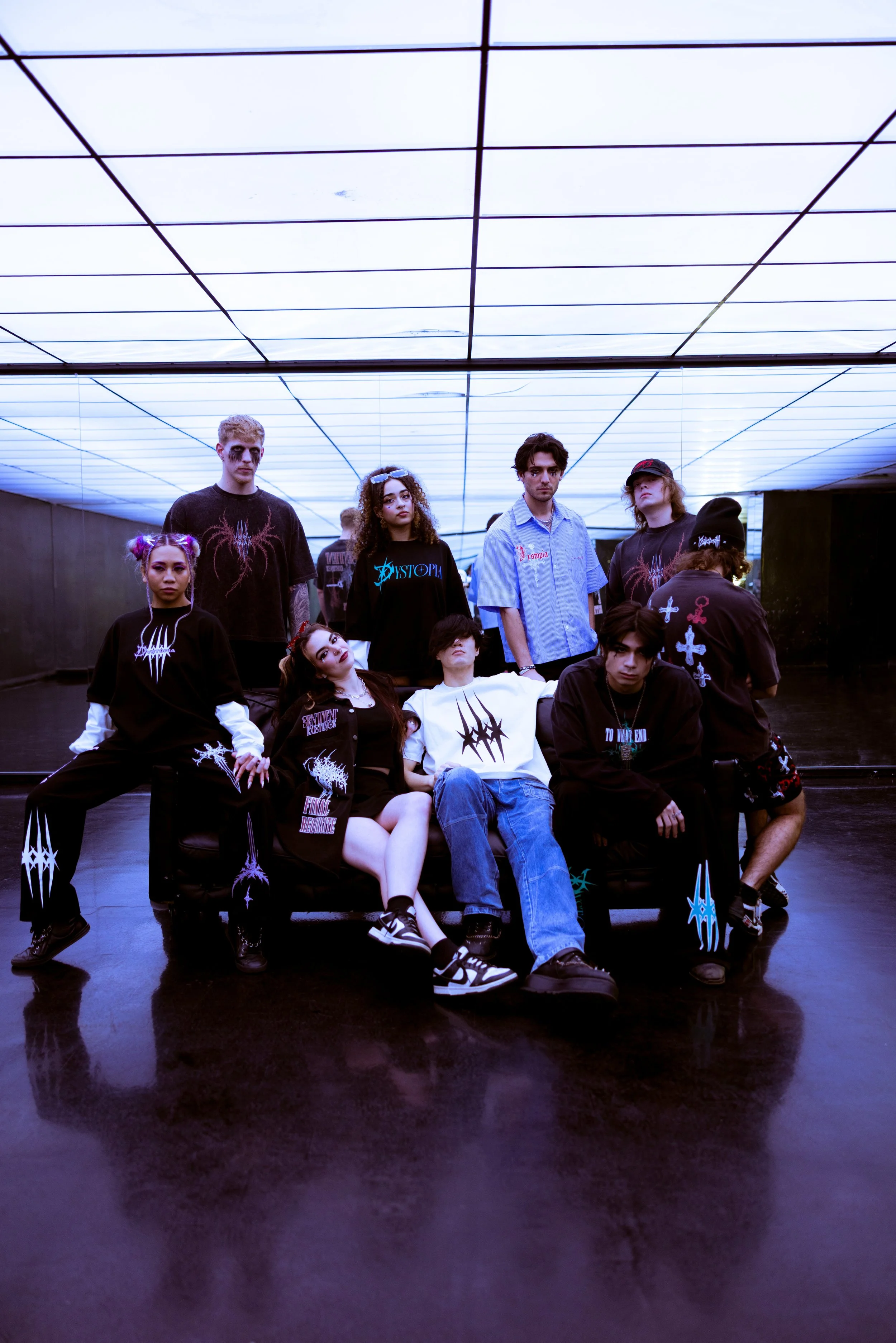 A group of young people in dark clothing with graphic designs, some with makeup, posing in a modern, illuminated indoor space with a reflective floor and a ceiling with a grid pattern.