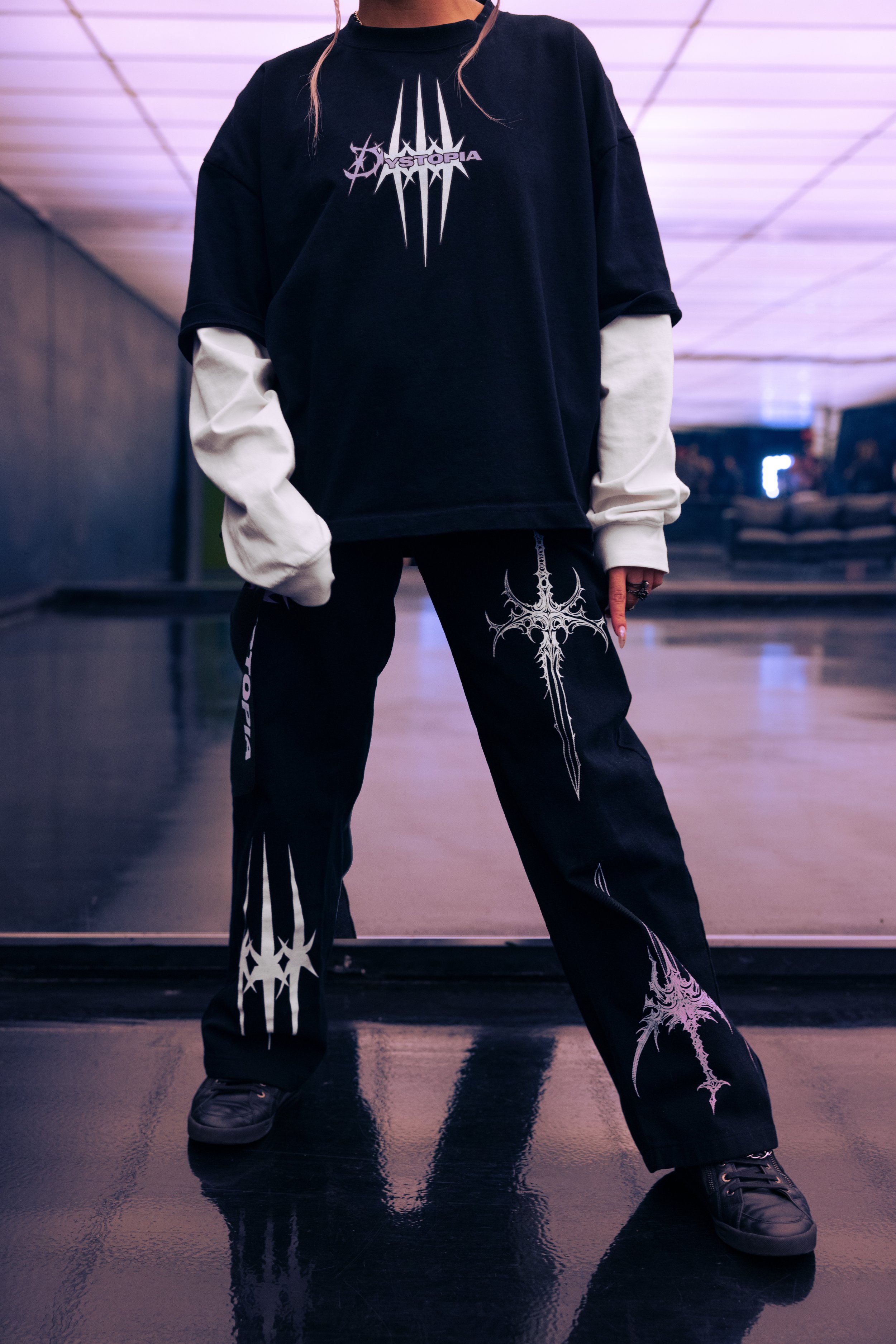 Person wearing a black oversized t-shirt with a white graphic, layered over a white long-sleeve shirt, black pants with white graphic designs, and black shoes, standing in a dimly lit indoor space with reflective flooring.
