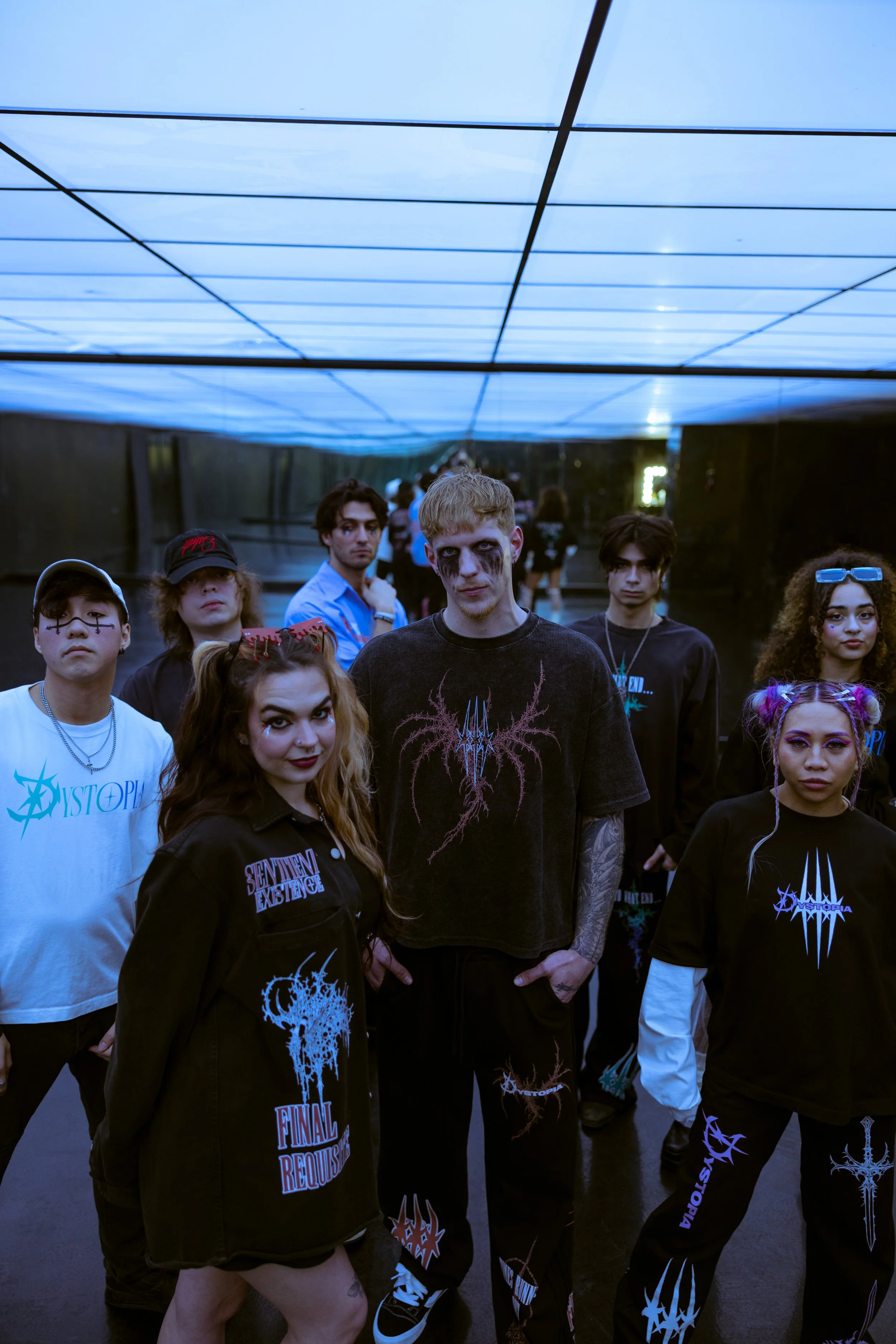 A group of young people dressed in goth clothing with dark makeup, standing inside a modern corridor with a blue glowing ceiling.