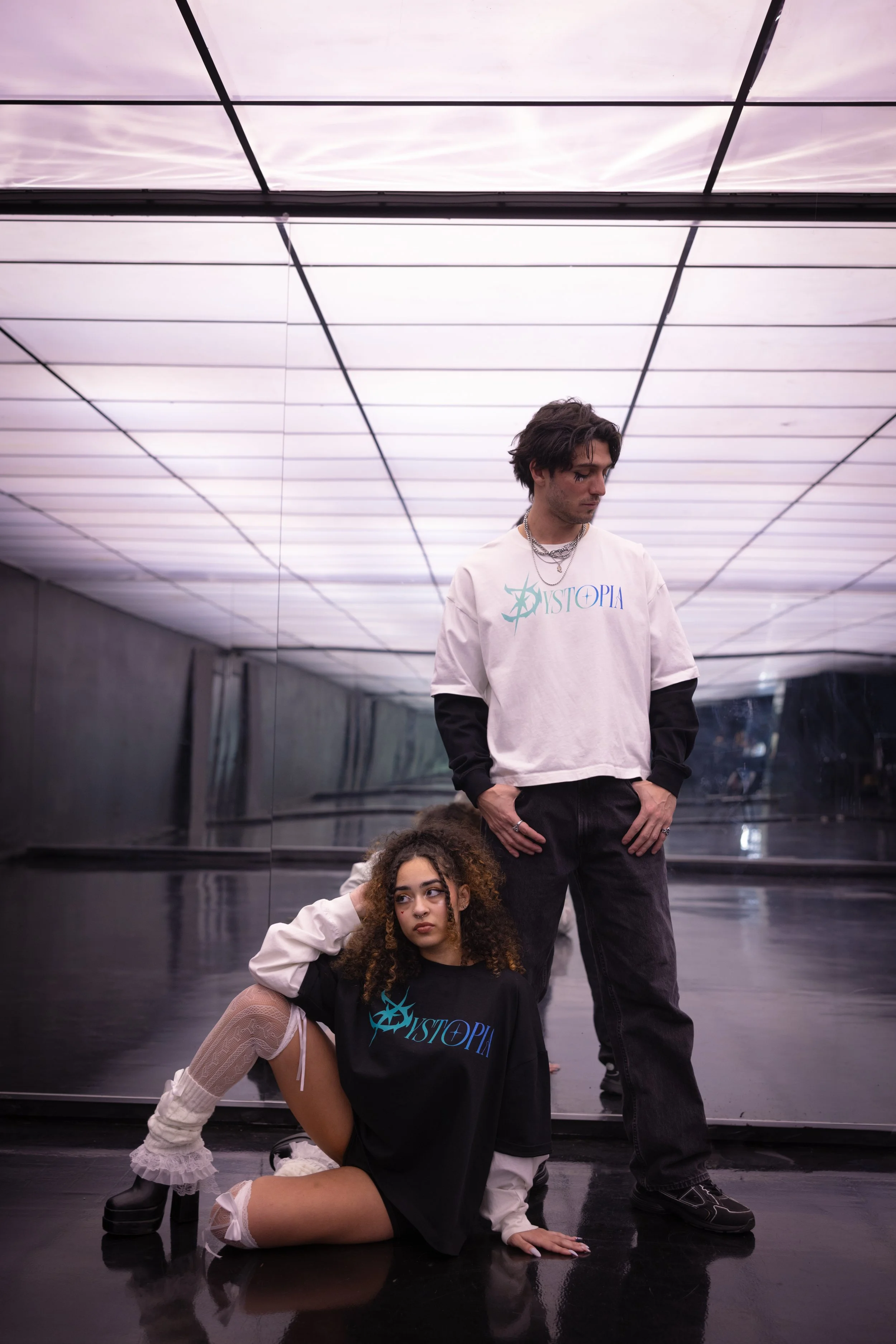 A young man and woman pose in front of a reflective mirrored wall in an illuminated room. The man stands with his hands in his pockets, looking to the side, while the woman sits on the floor with one hand on her head, looking ahead.