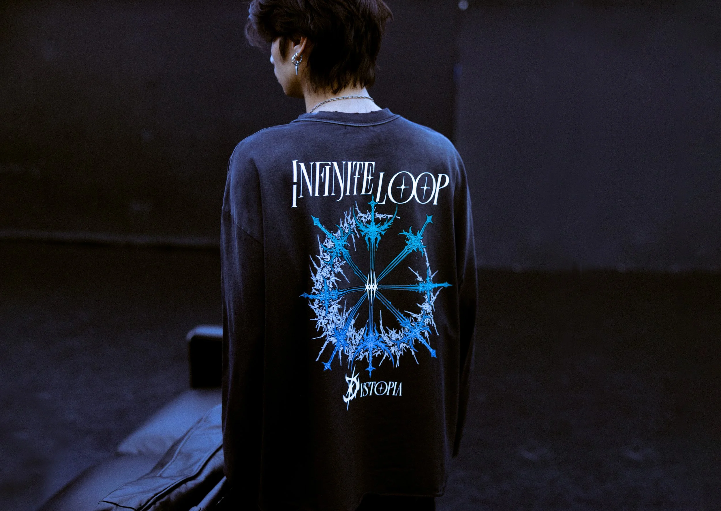 Person with short dark hair wearing a black long-sleeve shirt with a graphic and text on back, standing in a dark room.