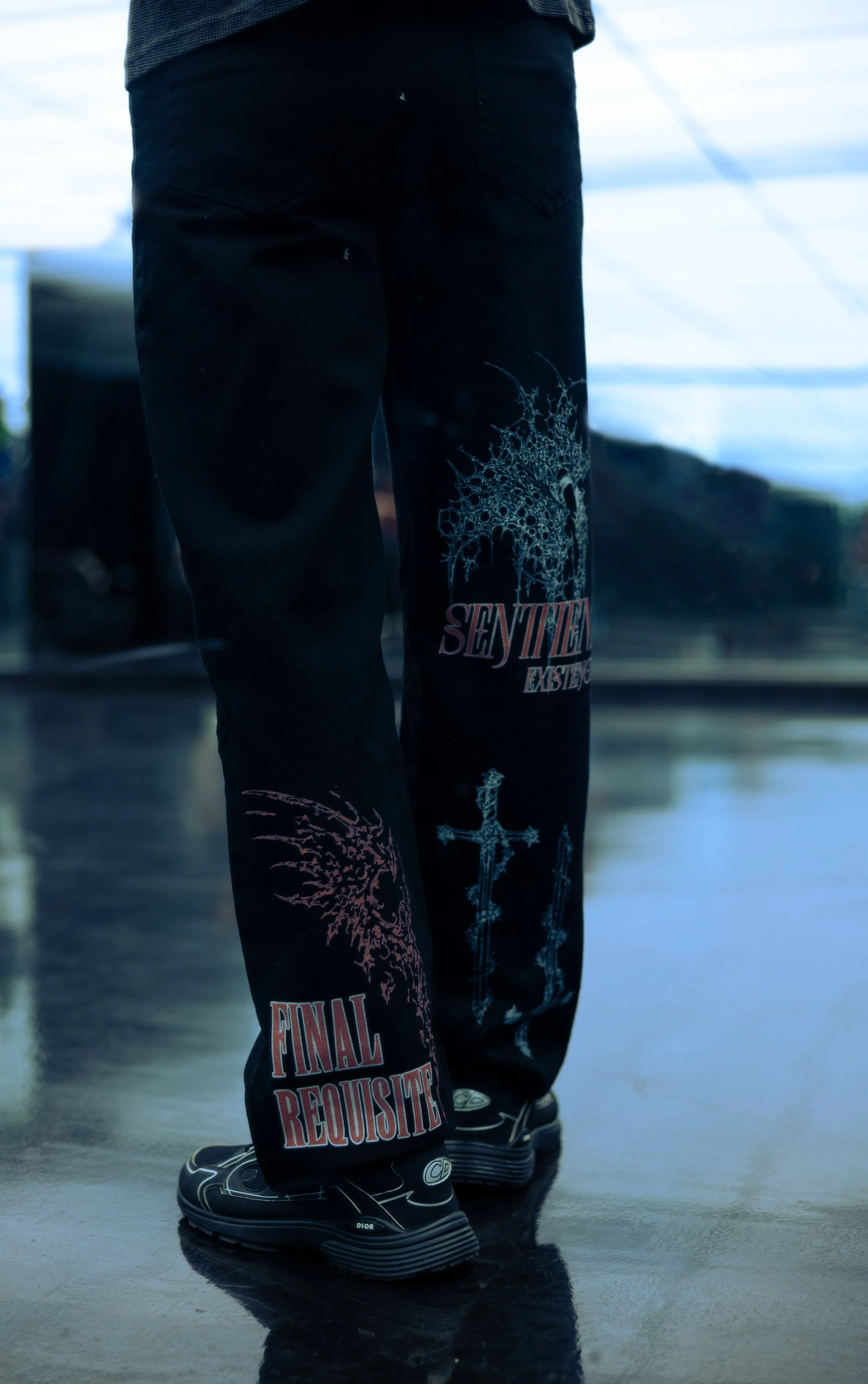 Person standing indoors wearing black pants with band logos and black sneakers, with a reflective floor and a window in the background.