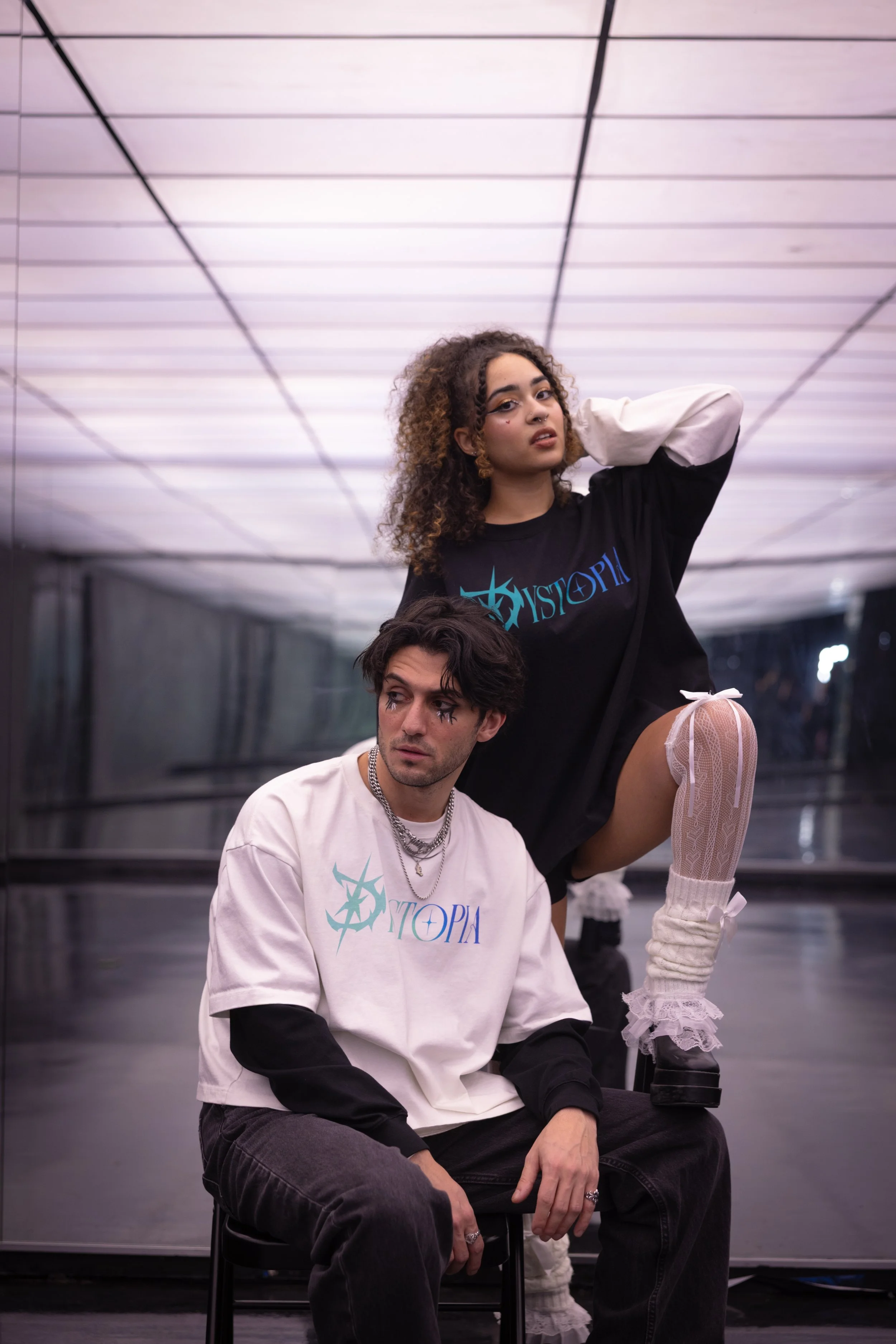 A young woman with curly hair wearing a black t-shirt and white knee-high stockings with lace and bows, posing with one leg raised on a man’s lap. The man has dark hair, is wearing a white t-shirt, black pants, and layered necklaces. They are in a mo