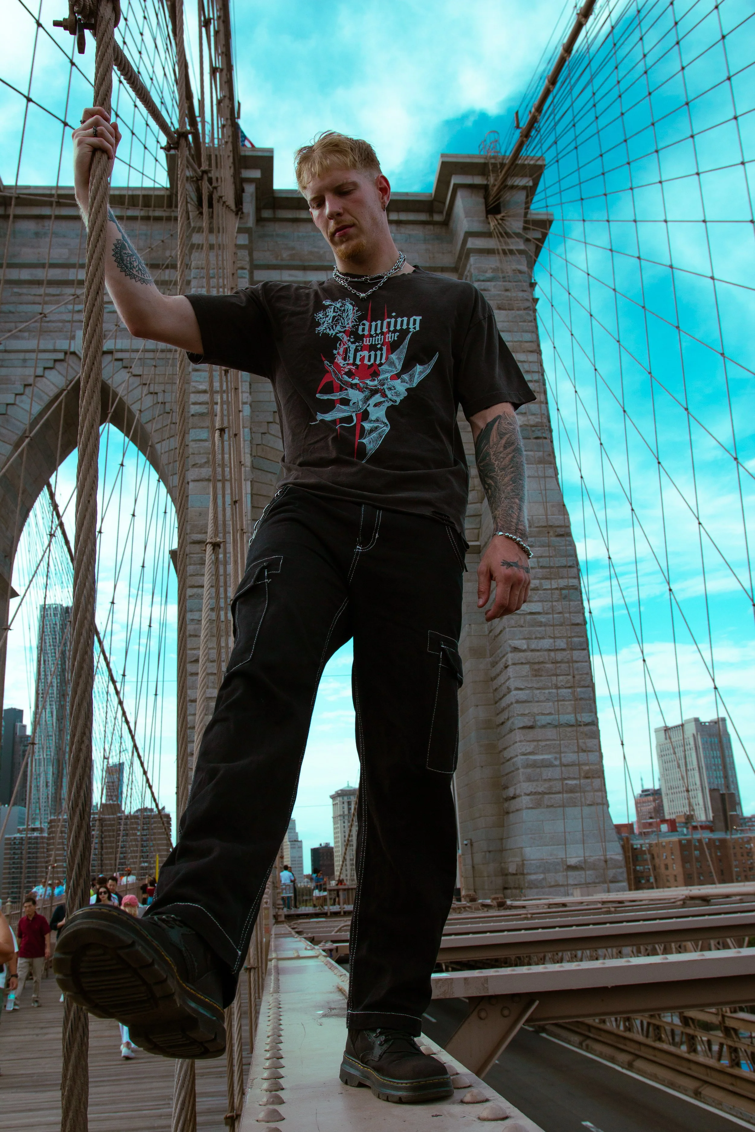 A young man is standing on the Brooklyn Bridge in New York City, holding a rope with one hand. He is wearing a black T-shirt with a graphic design and text, black cargo pants, and black boots. The city skyline is visible in the background under a par
