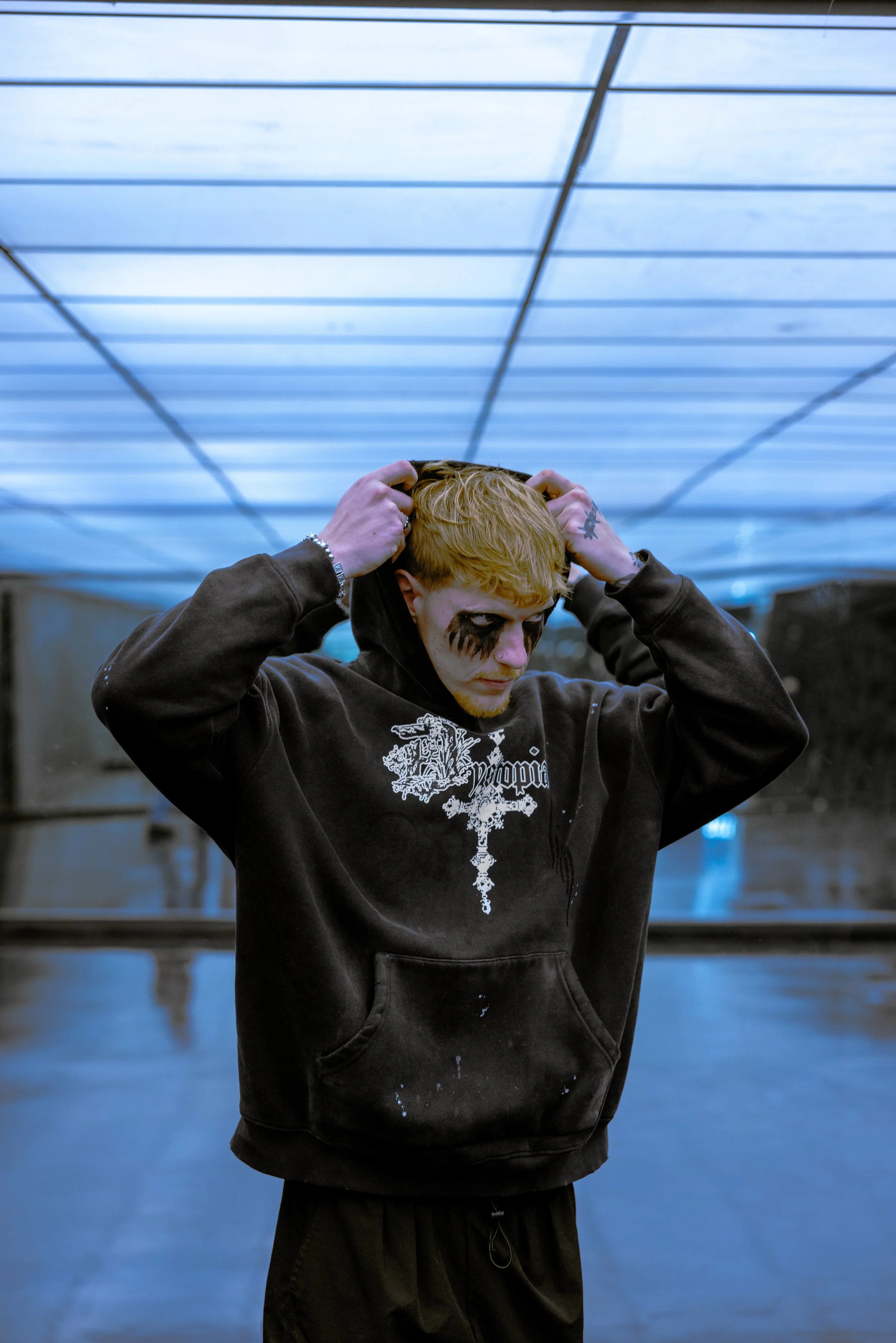 A young person with blond hair and face paint is adjusting their black hoodie indoors under a reflective ceiling.