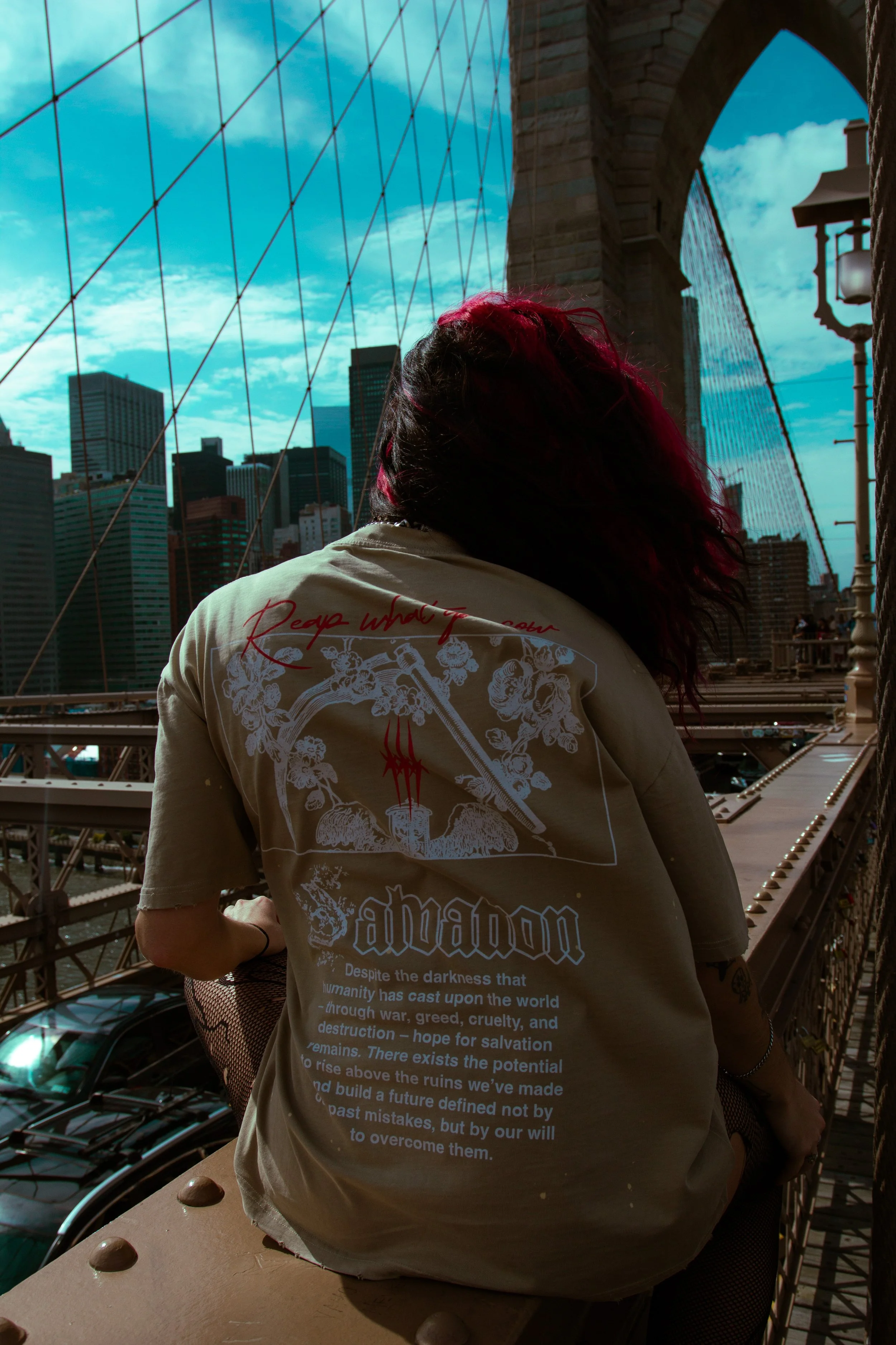 Person with wavy hair and red highlights sitting on the edge of the Brooklyn Bridge, overlooking the Manhattan skyline in New York City with skyscrapers and cranes.