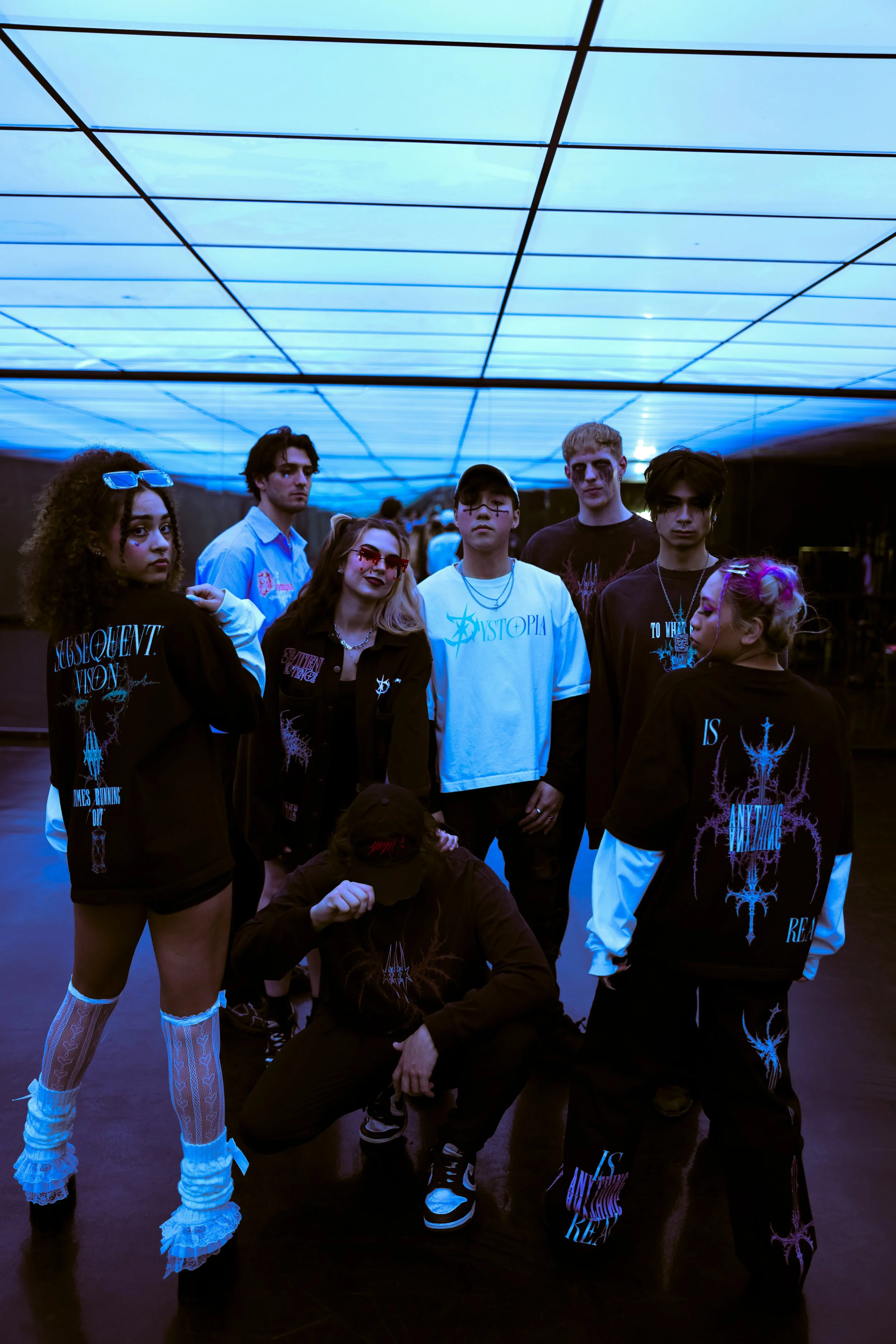 Group of young people posing in a dark, modern room with a glowing blue ceiling, dressed in black and white streetwear with gothic and alternative fashion styles.