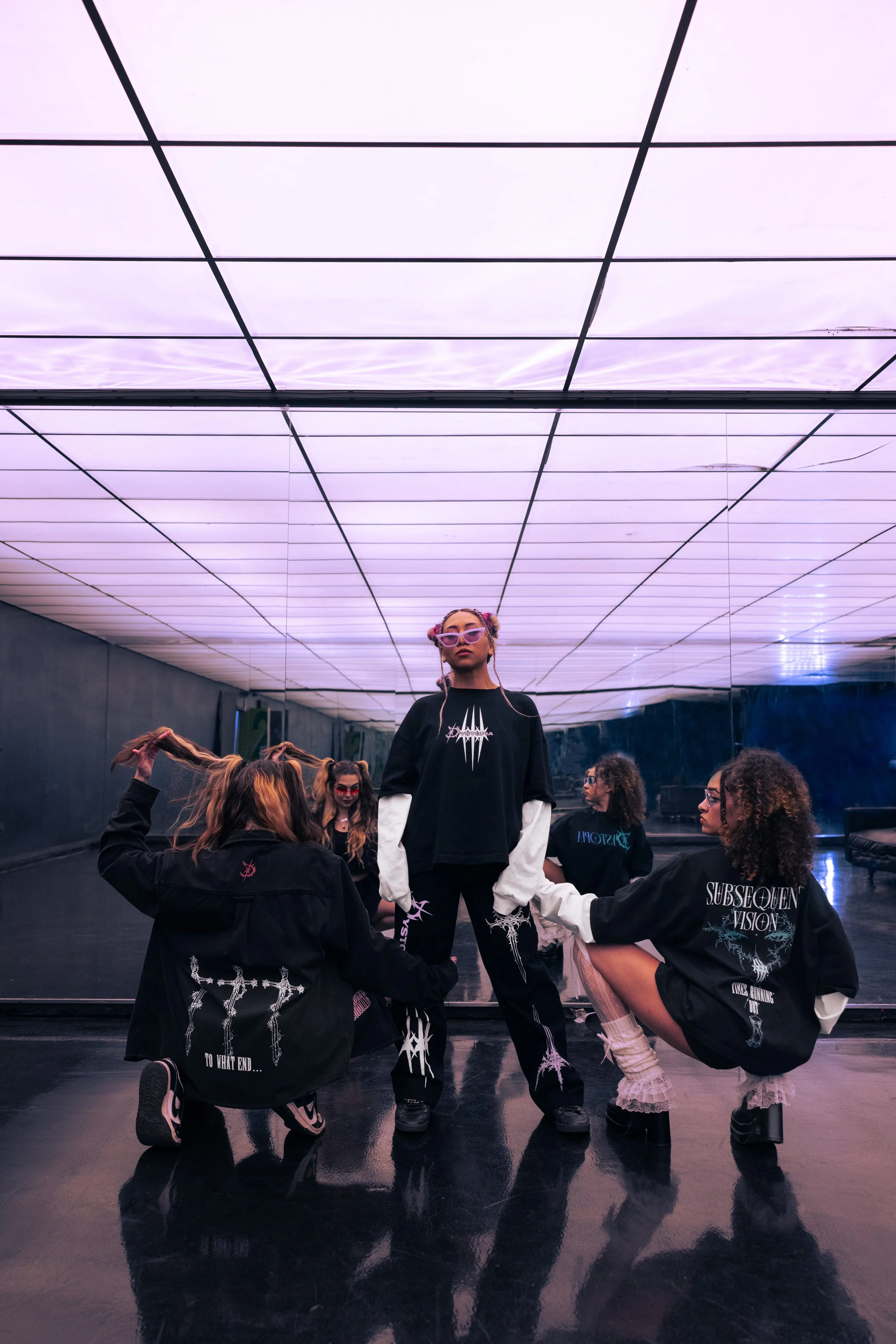 A group of young women with edgy fashion style, black graphic sweatshirts, and headphones, posing in a dimly-lit dance studio with futuristic purple ceiling lights.