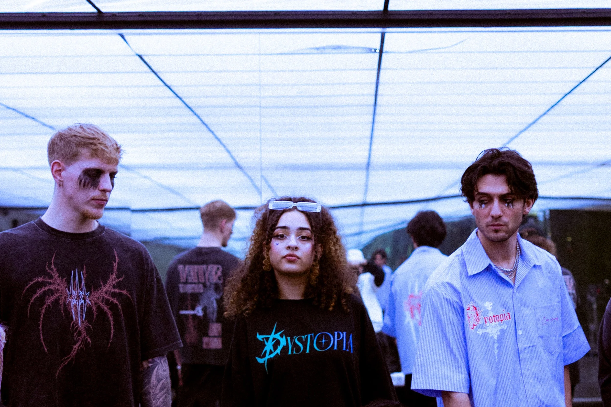 Group of young people at an outdoor event under a canopy, with the central female wearing a black shirt with blue text and two young men in casual clothing on either side.