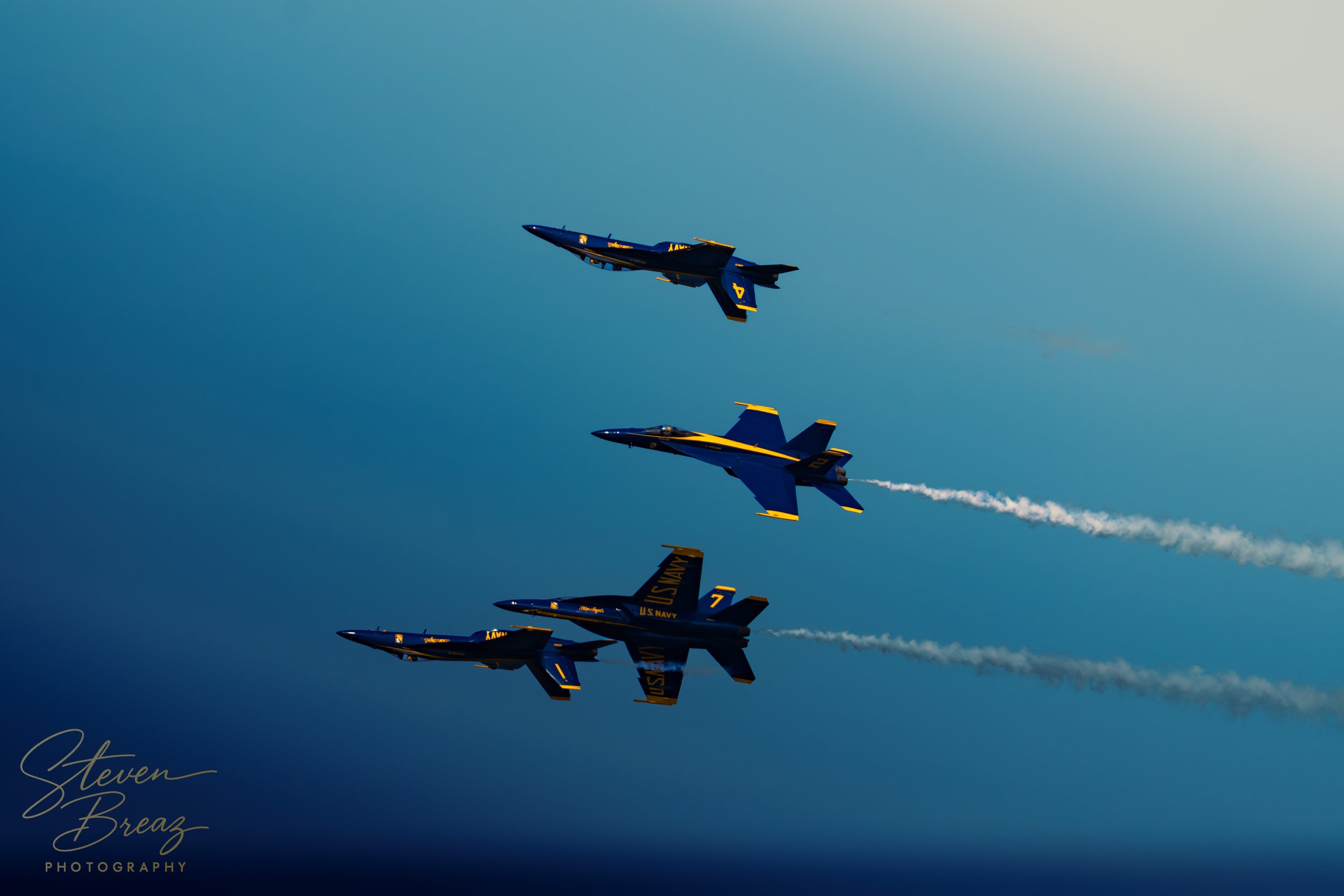 Four US Navy fighter jets flying in close formation against a blue sky, leaving white smoke trails behind them.