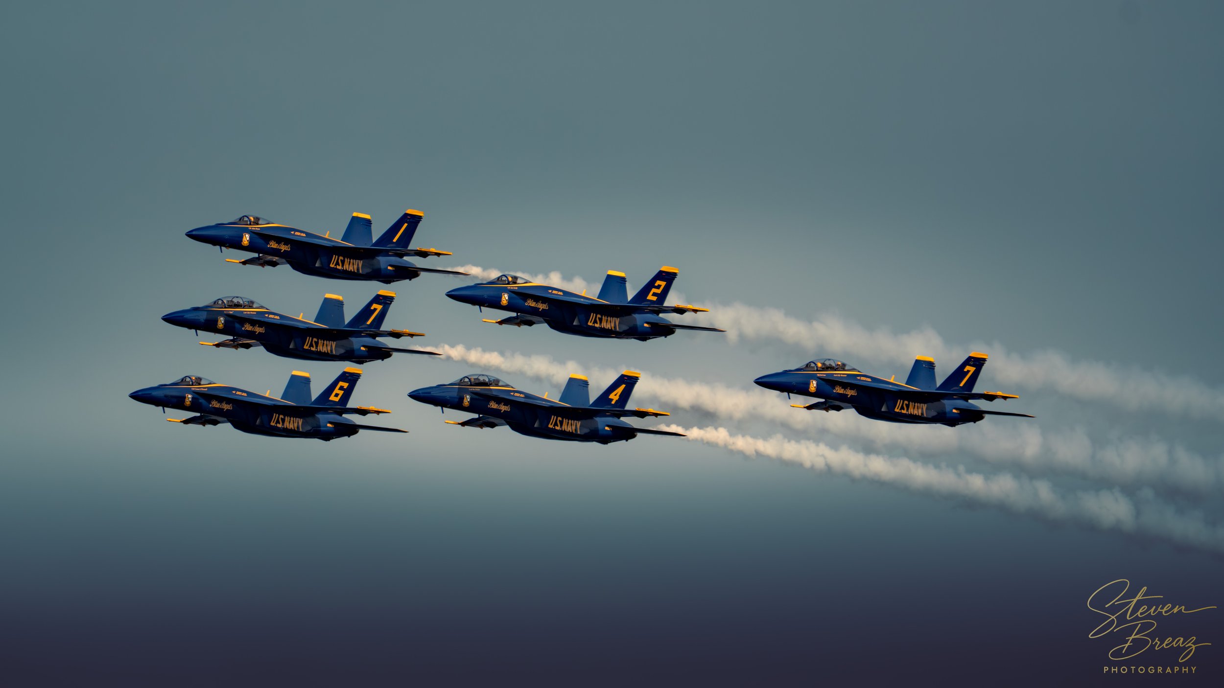 Six U.S. Navy blue fighter jets flying in tight formation, leaving white smoke trails against a cloudy sky.