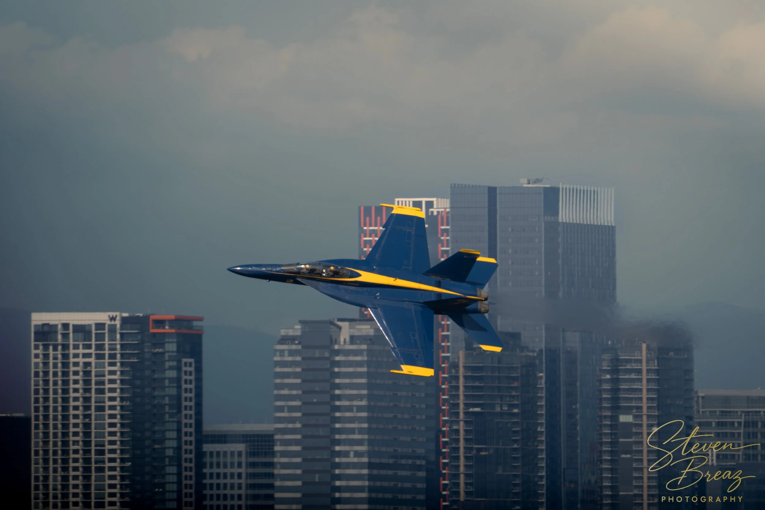 A blue and yellow fighter jet flying over a city skyline with tall buildings and a cloudy sky.