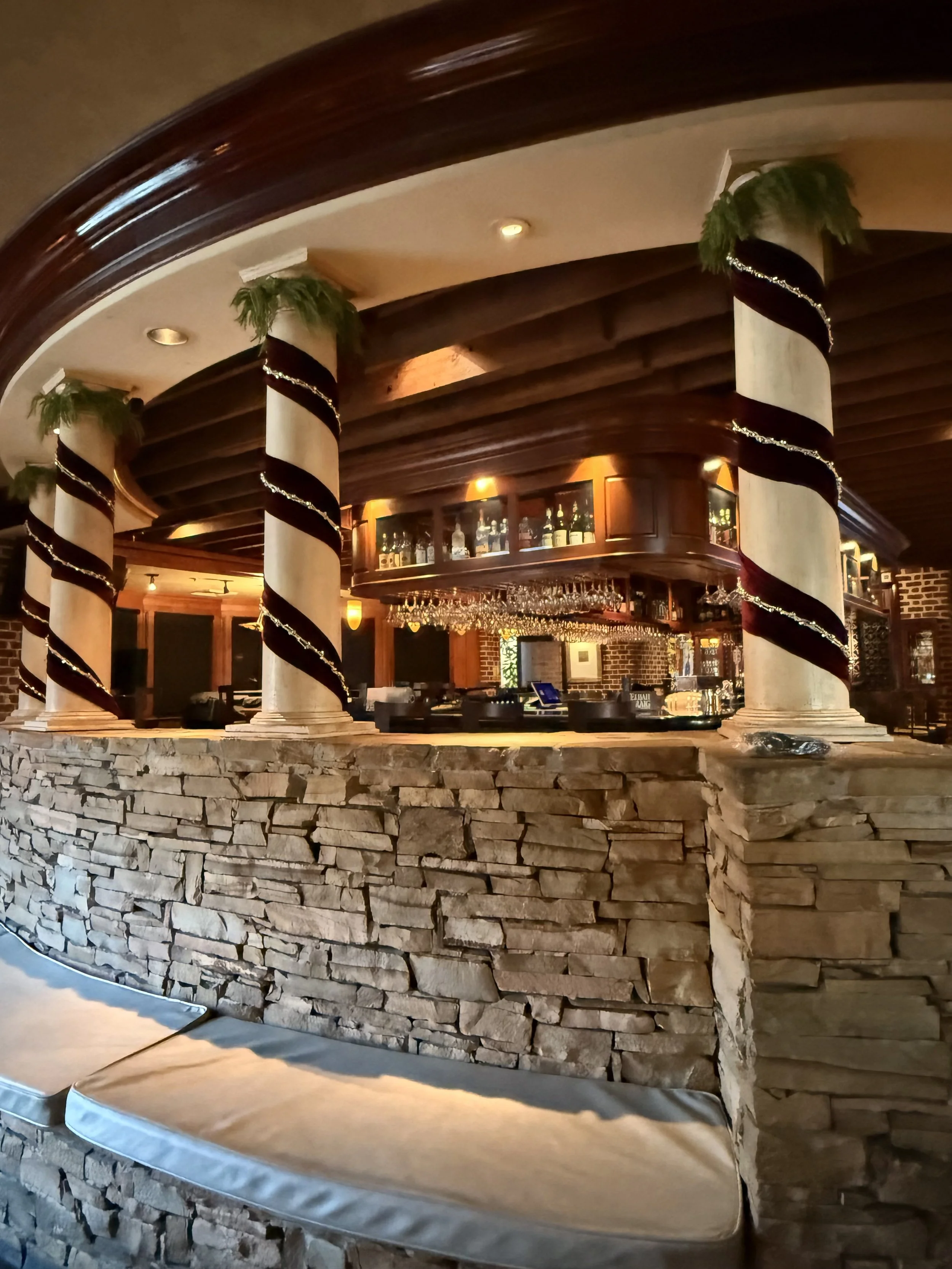 Indoor room decorated for Christmas with 4 columns wrapped in burgundy velvet and gold beads. Bench seating with stacked stone and and bar setting with glassware in background.
