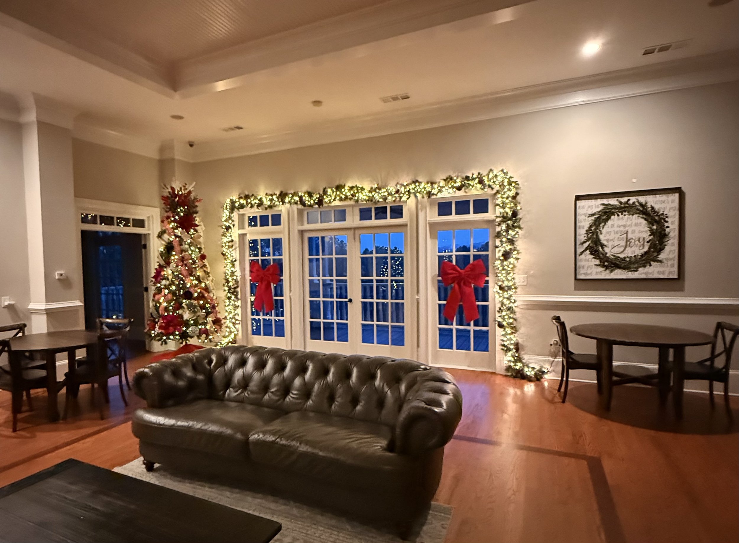 Large indoor living room with leather couch, tables with chairs, back window decorated with garland, lights and red bows. Christmas tree decorated with red and white ornaments. Large picture on wall of a wreath.