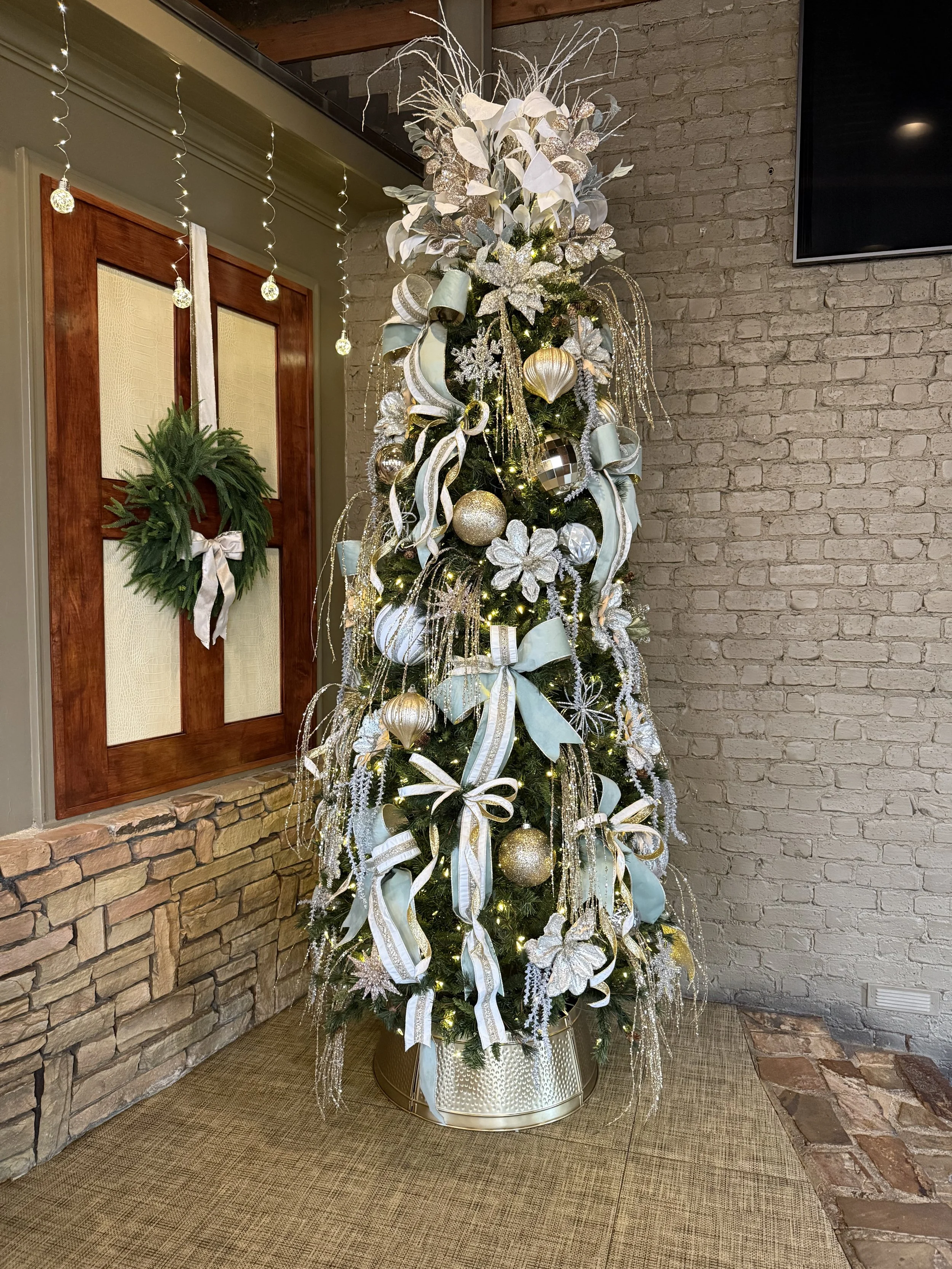 Decorated Christmas tree with white and silver ornaments, sage green ribbons, and lights, topped with silver branches, standing on a gold tree collar with a green wreath with a bow hanging on adjacent wall..