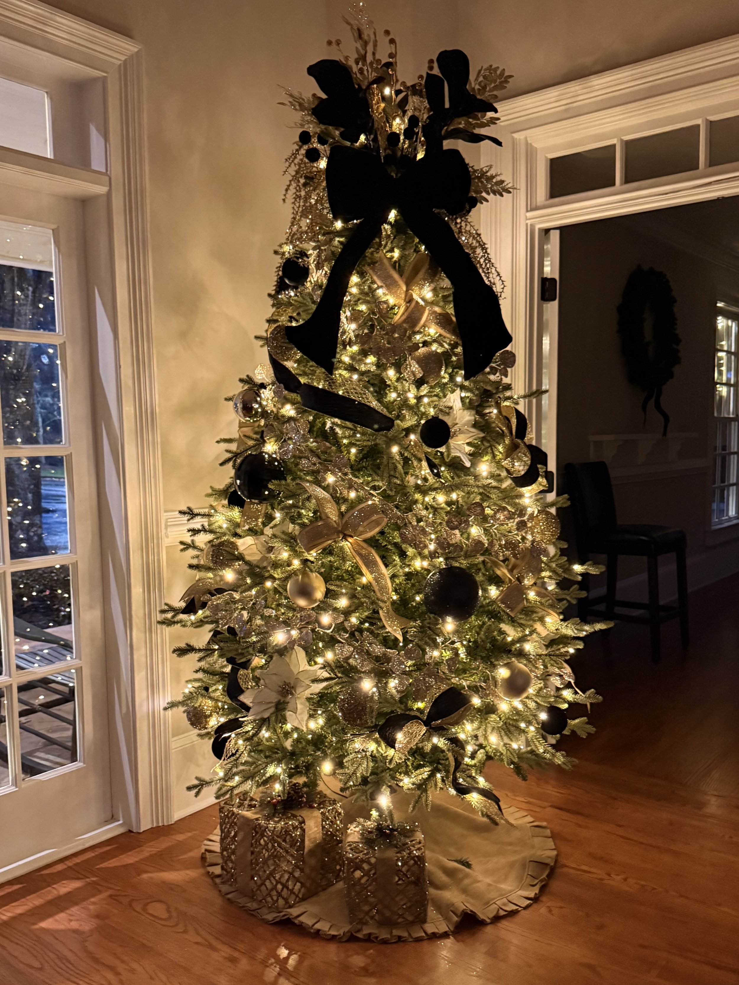 Indoor Christmas tree with black ribbon bow, gold, black and white ornaments in the corner of the room, next to a window and gold gifts underneath.