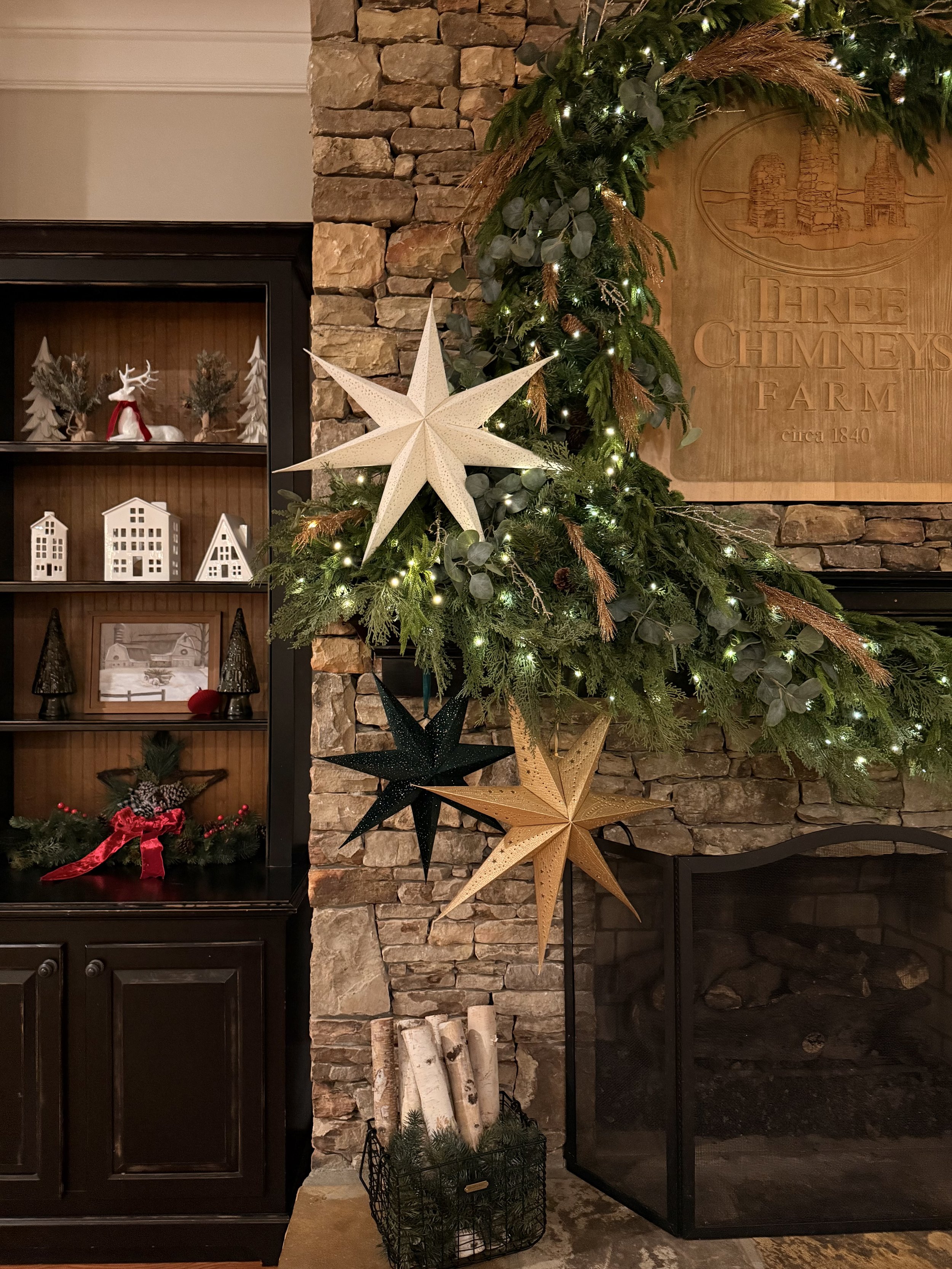 Stacked stone mantle and fireplace with green garland lights, white, green and gold stars next to a cabinet with festive decor on shelves.