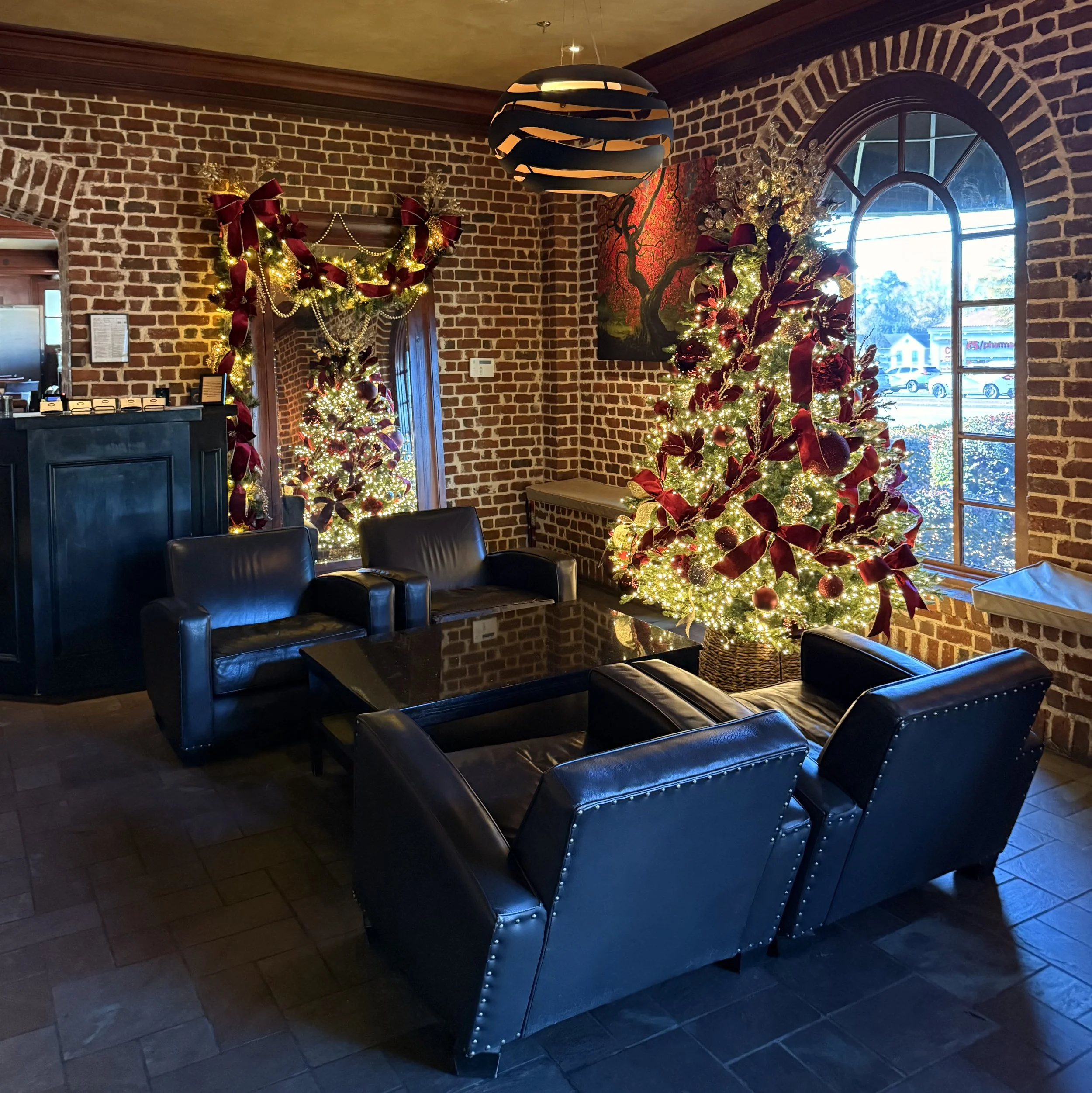 Restaurant lobby with brick walls, 4 leather chairs, hostess stand, christmas tree in front of front facing window, Tree and large mirror decorated with burgundy velvet ribbon, champagne ornaments,  