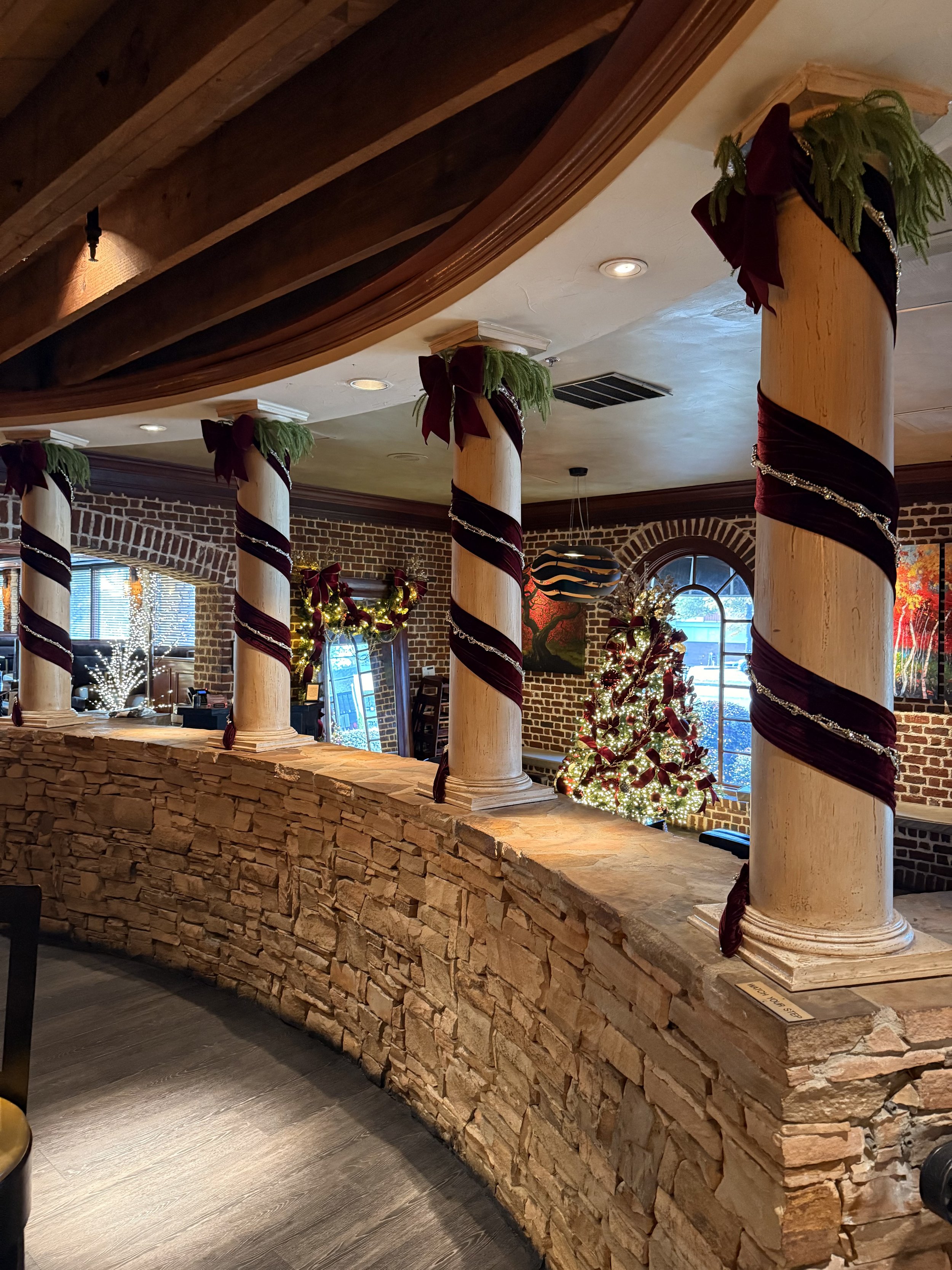 Indoor room decorated for Christmas with 4 columns wrapped in burgundy velvet and gold beads. Lobby Christmas tree decorated and arched window in the background.