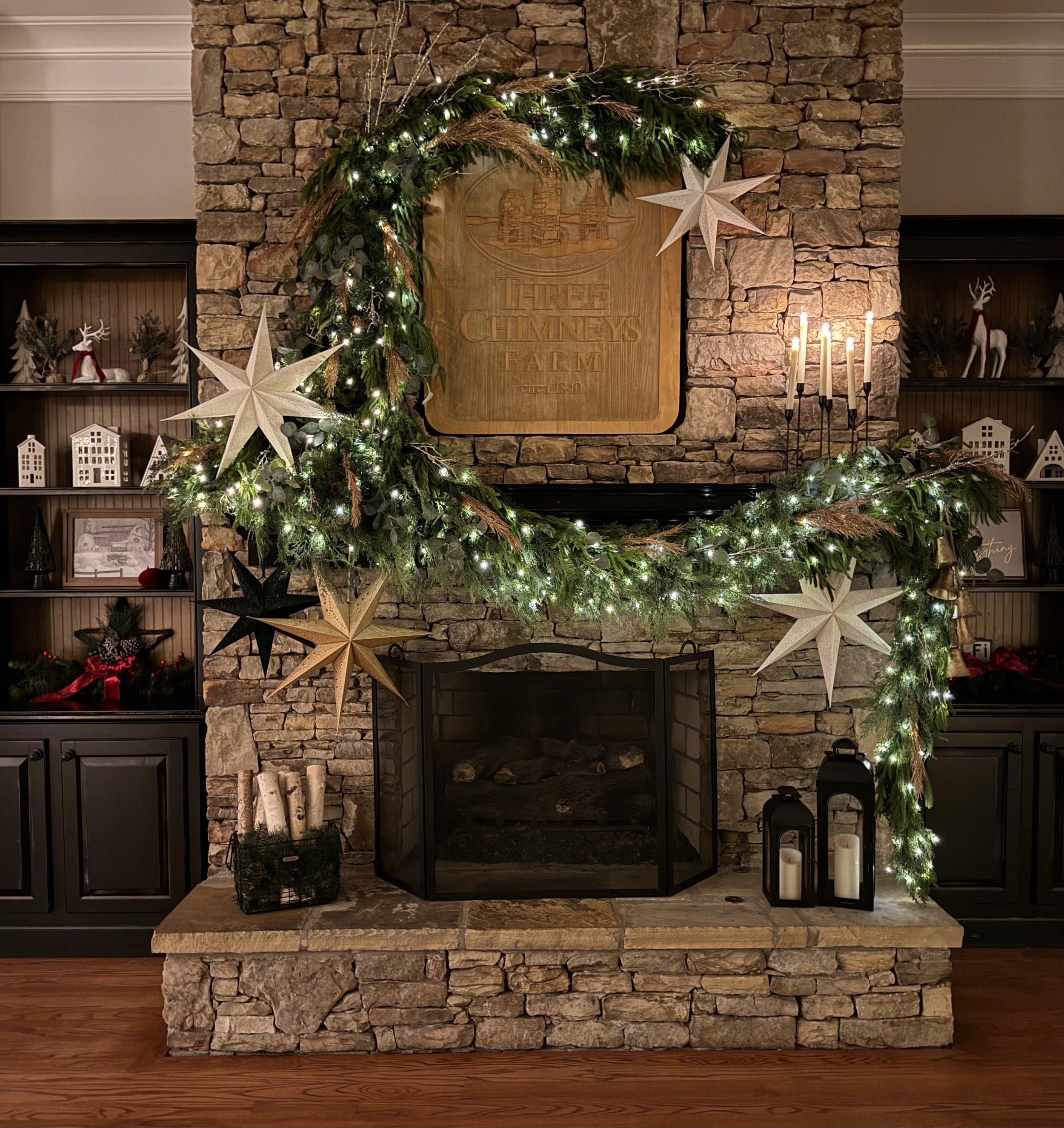 Decorated stone fireplace with a green Christmas garland, white stars, candles, and festive ornaments in a living room.