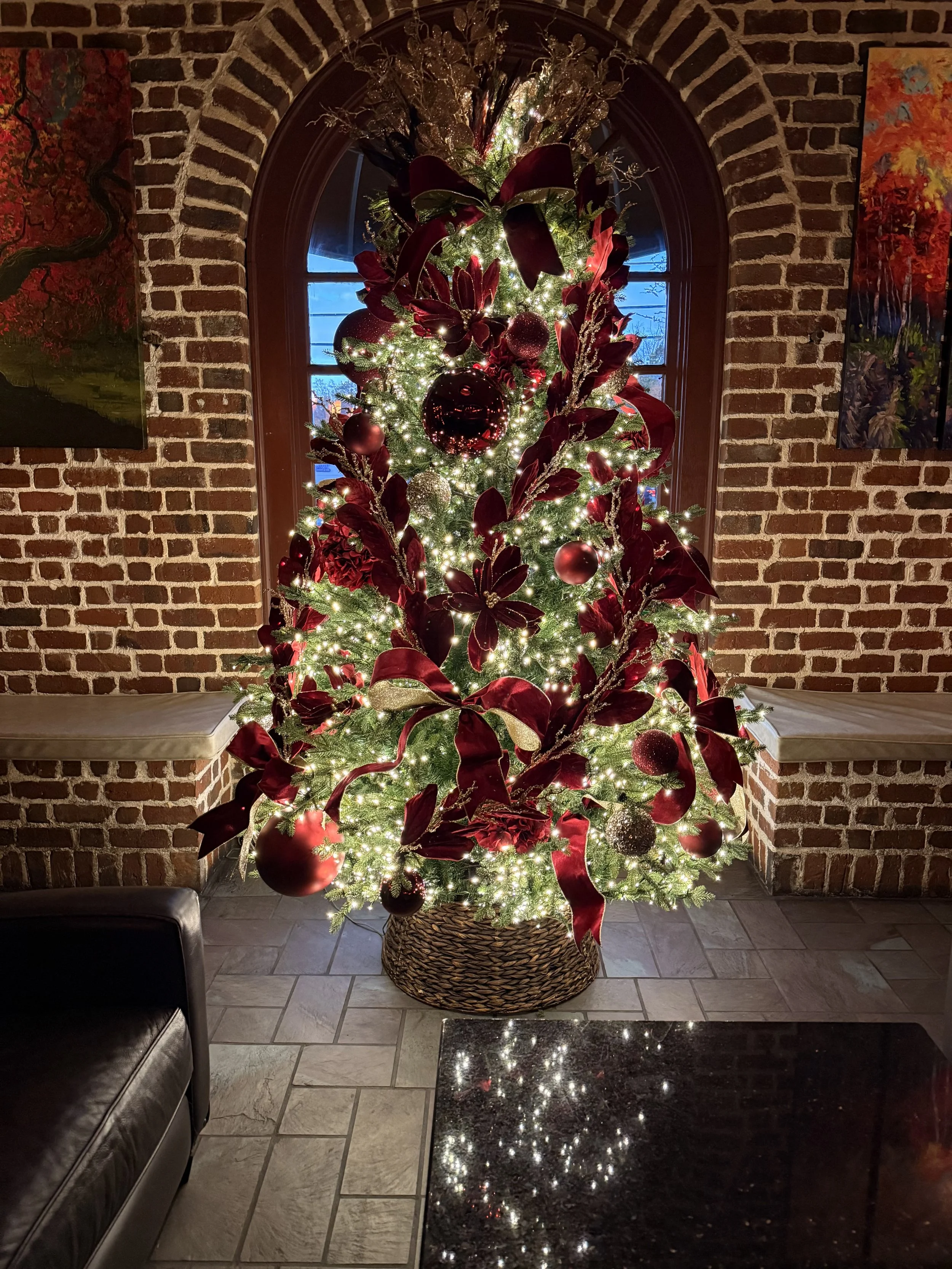 Decorated Christmas tree with red and gold ornaments, ribbon, and lights in front of a brick wall with a window.
