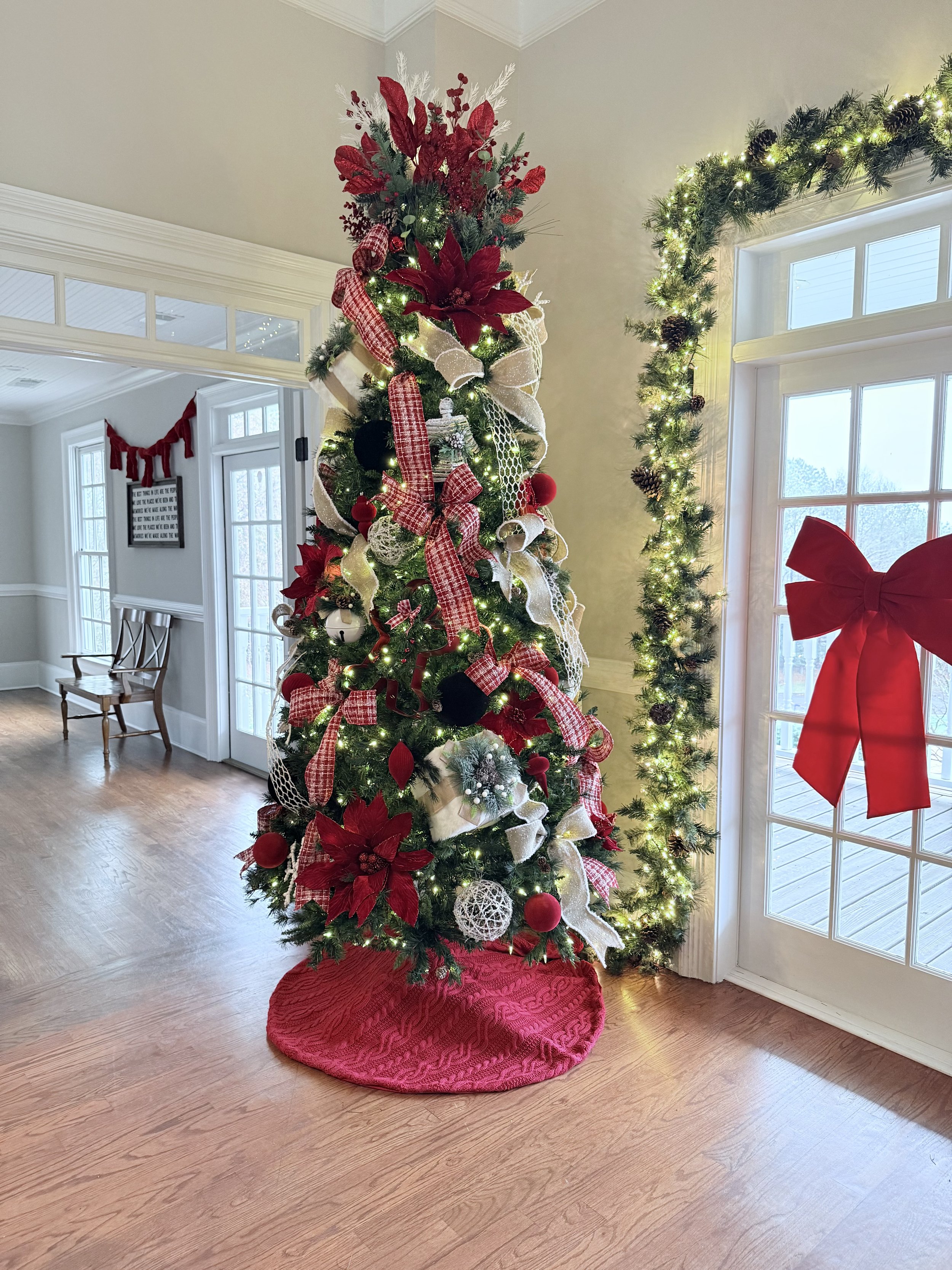 Decorated Christmas tree with red, white, and black ornaments, ribbons, and lights, placed in a room with a hardwood floor next to a door with a red bow on the window.