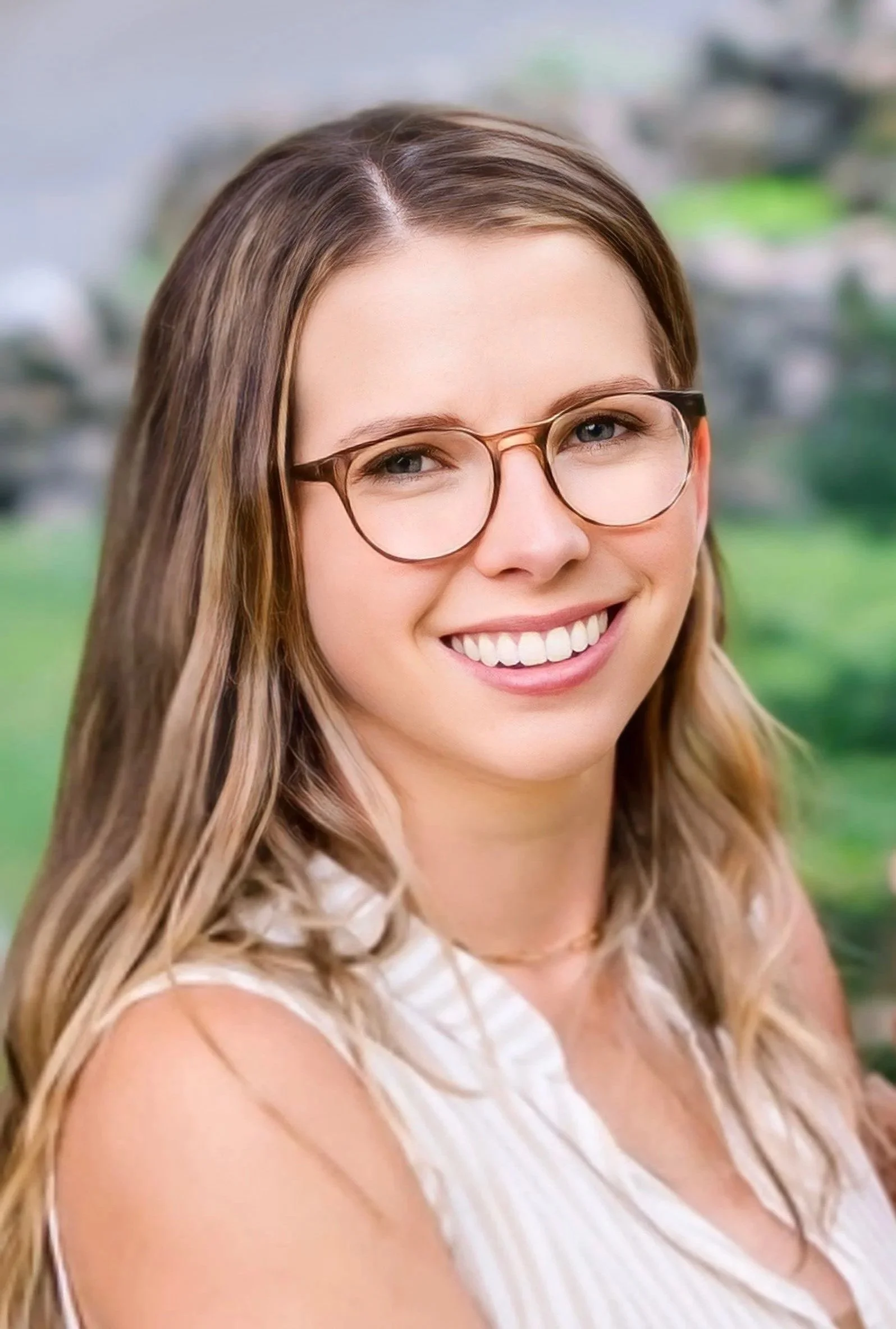A young woman with blonde hair, wearing glasses and a sleeveless top, smiling outdoors with a blurred green background.