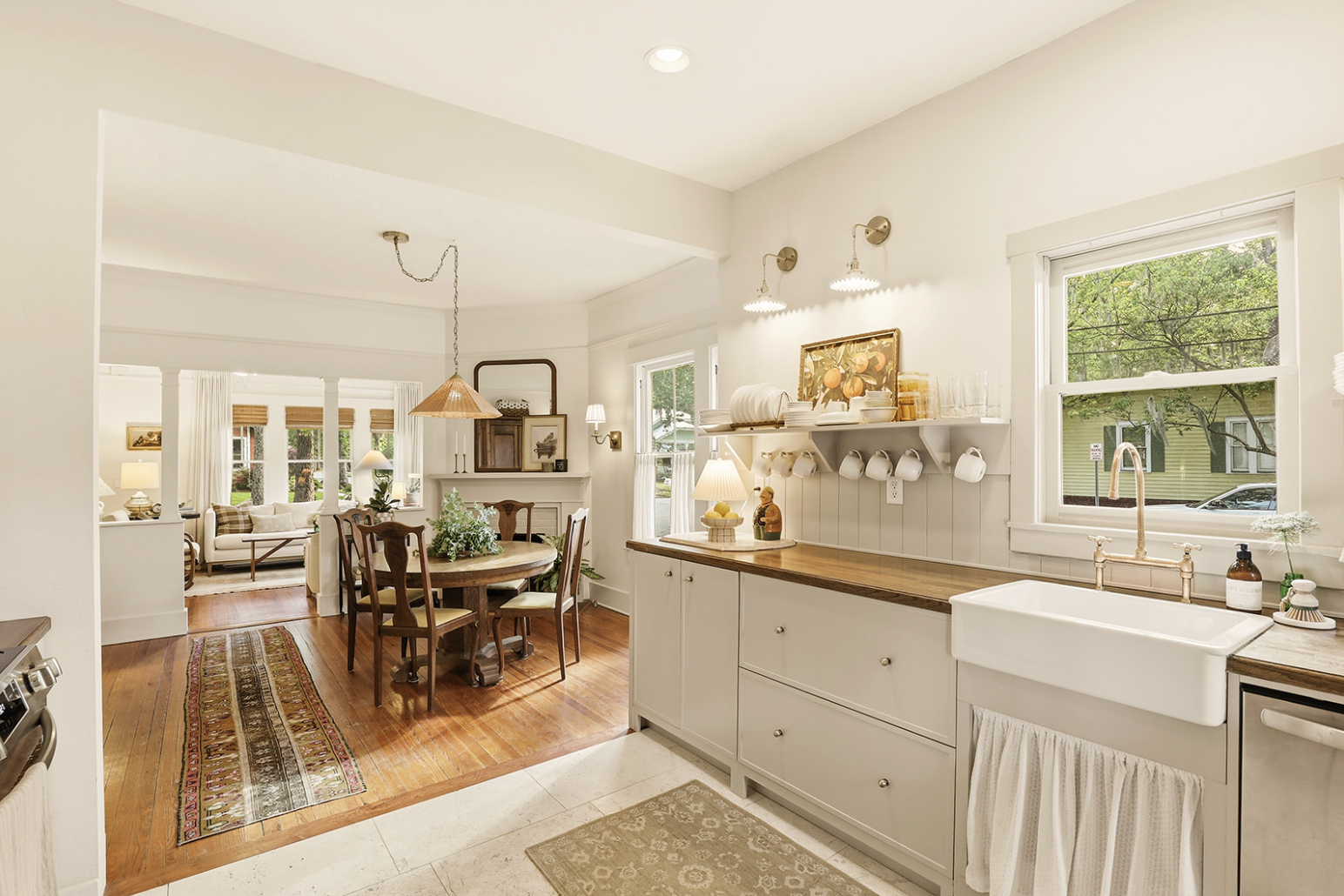 View of a cozy, well-lit kitchen connected to a dining and living area with large windows, wooden floors, white walls, a farmhouse sink, and decorative items.