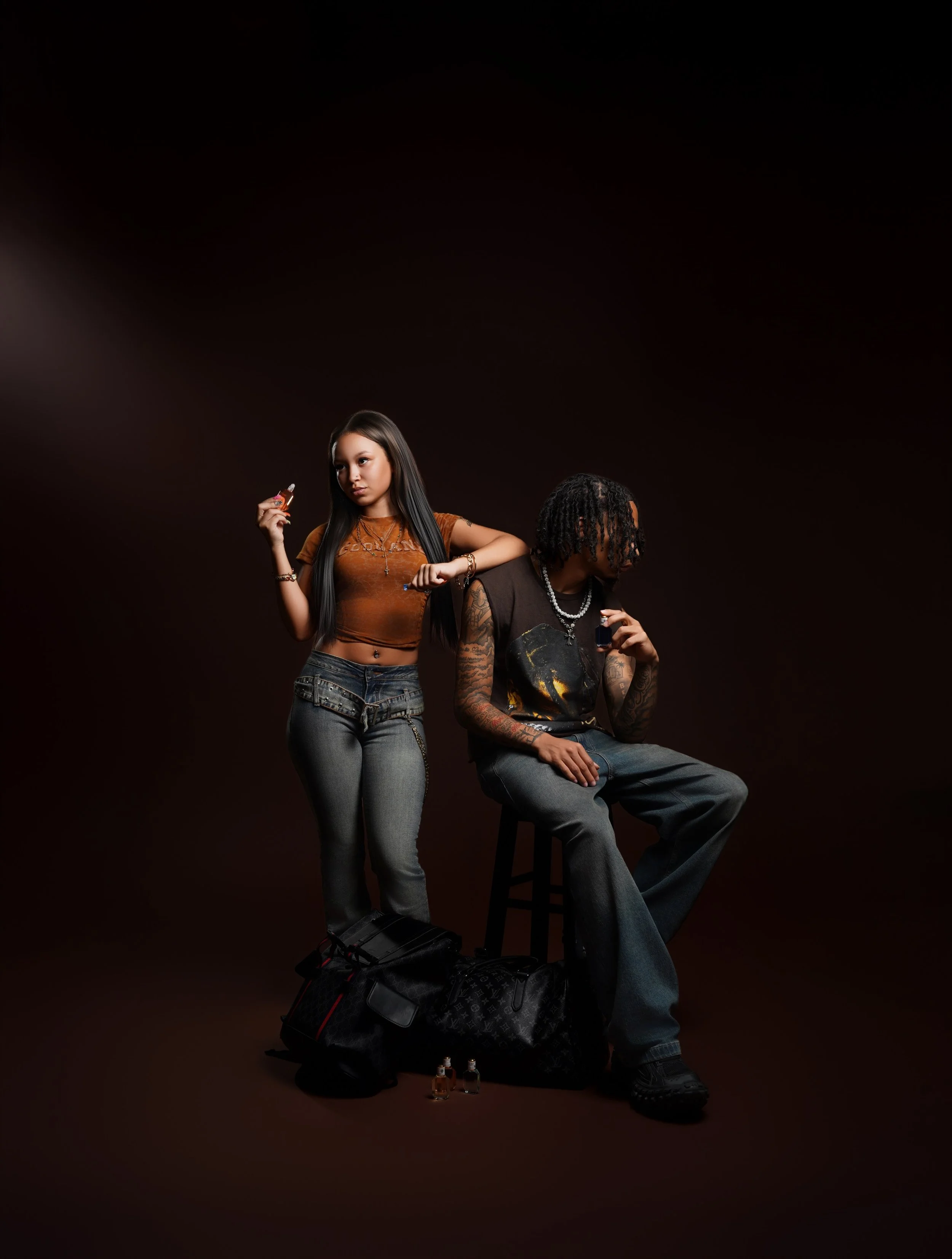 A young woman standing and a young man sitting on a stool, with a dark background and a duffle bag on the floor, surrounded by small perfume bottles.