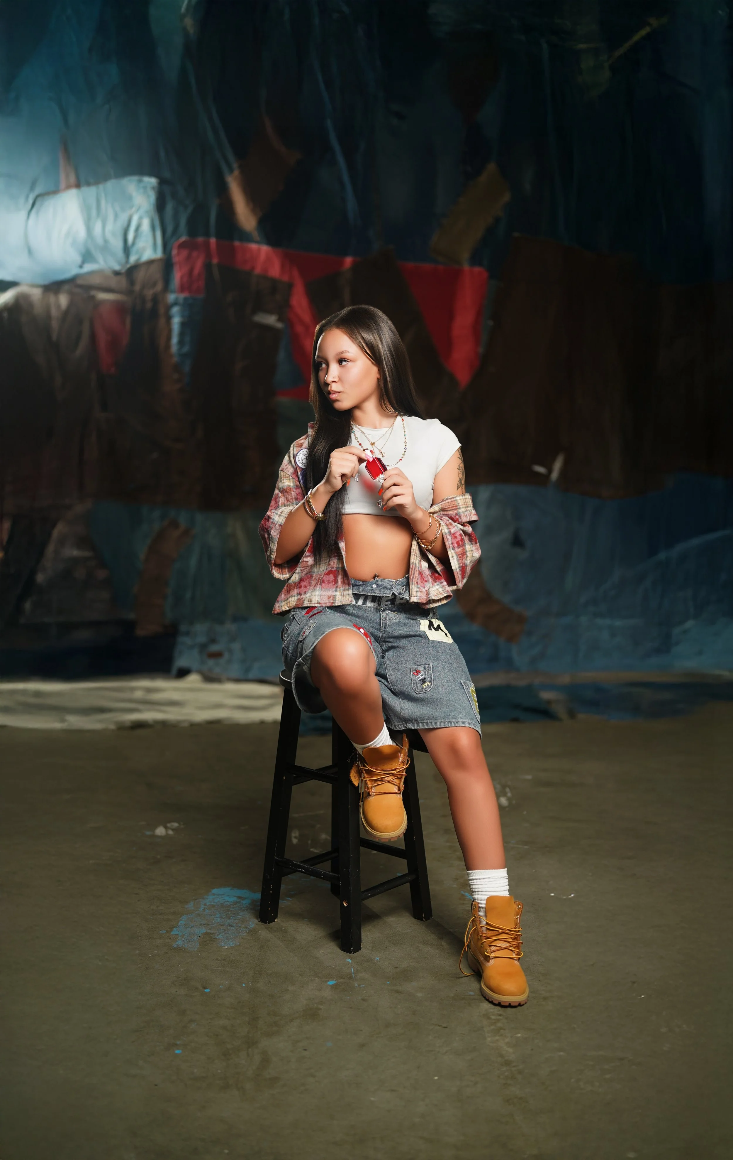 Young woman sitting on a black stool in a casual outfit with a plaid shirt, crop top, denim shorts, and tan boots, holding a red object, in an indoor space with a dark, painted backdrop.