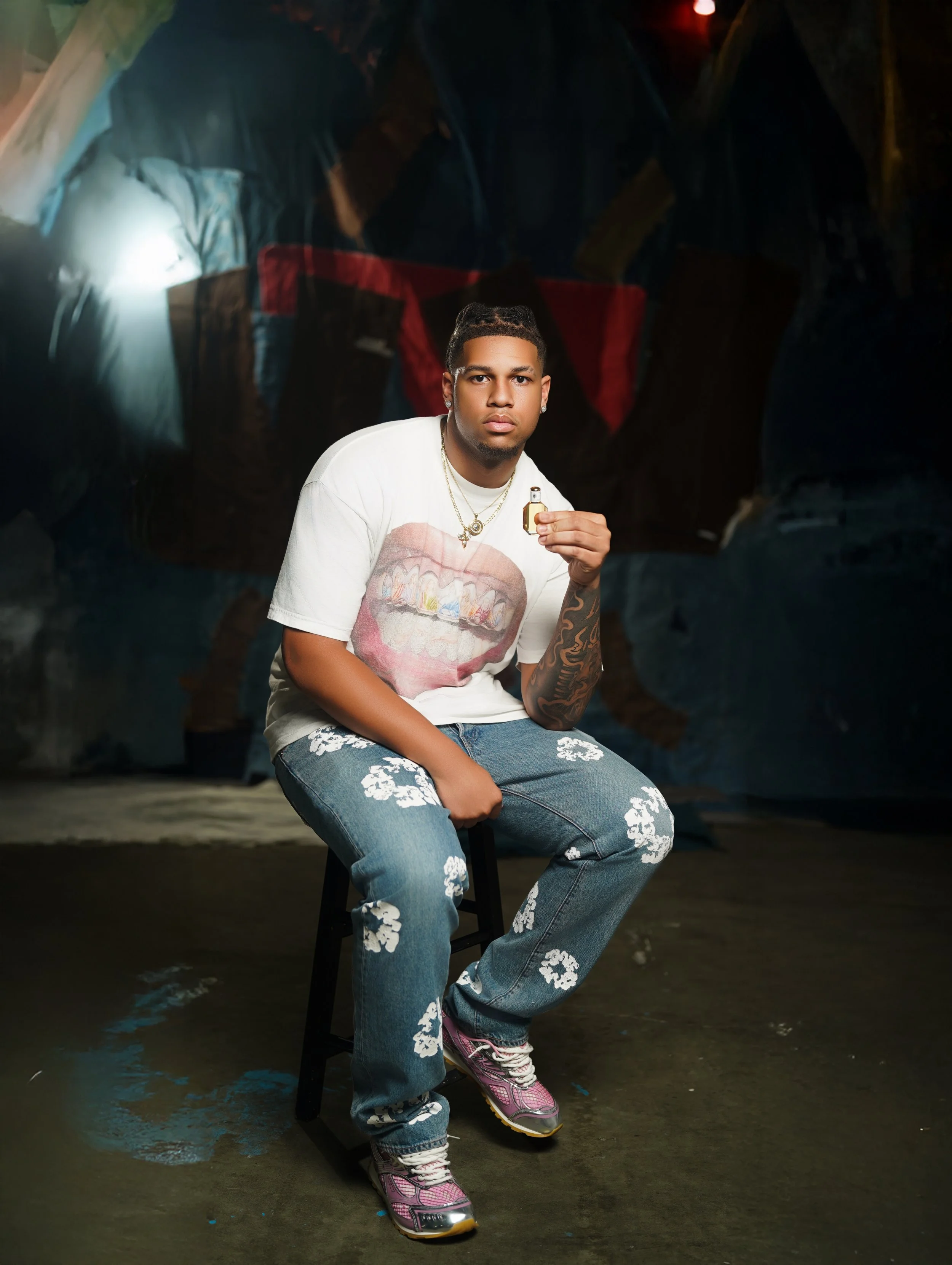 A young man with tattoos and jewelry sitting on a stool, holding a gold whistle, in front of a dark abstract painted background, wearing a white graphic t-shirt, floral jeans, and pink sneakers.