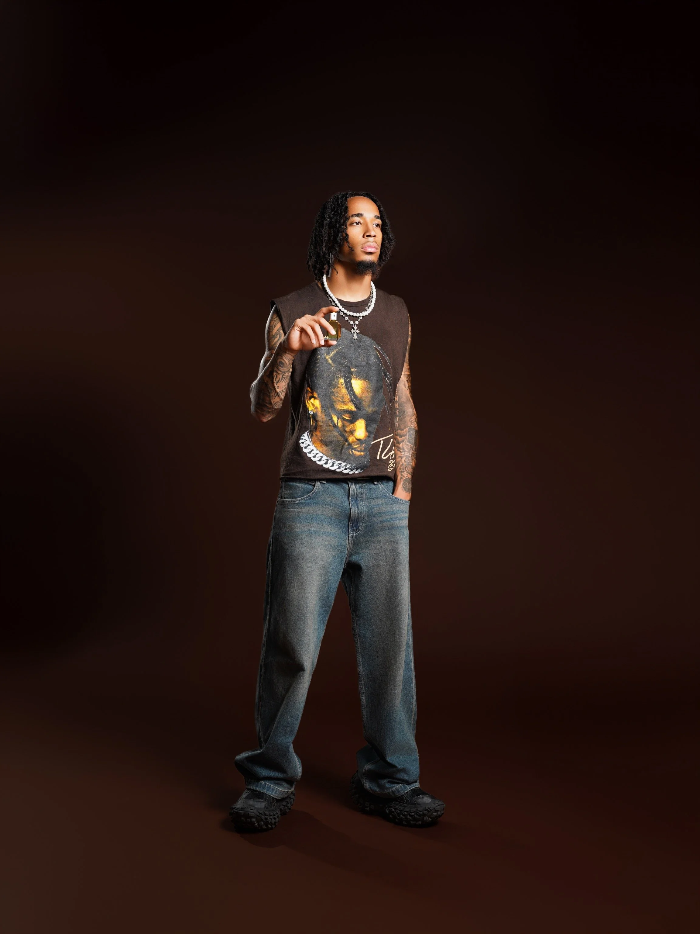 A young man with dark curly hair and tattoos, wearing a graphic T-shirt, jeans, and sneakers, holds a small bottle, standing against a dark brown background.