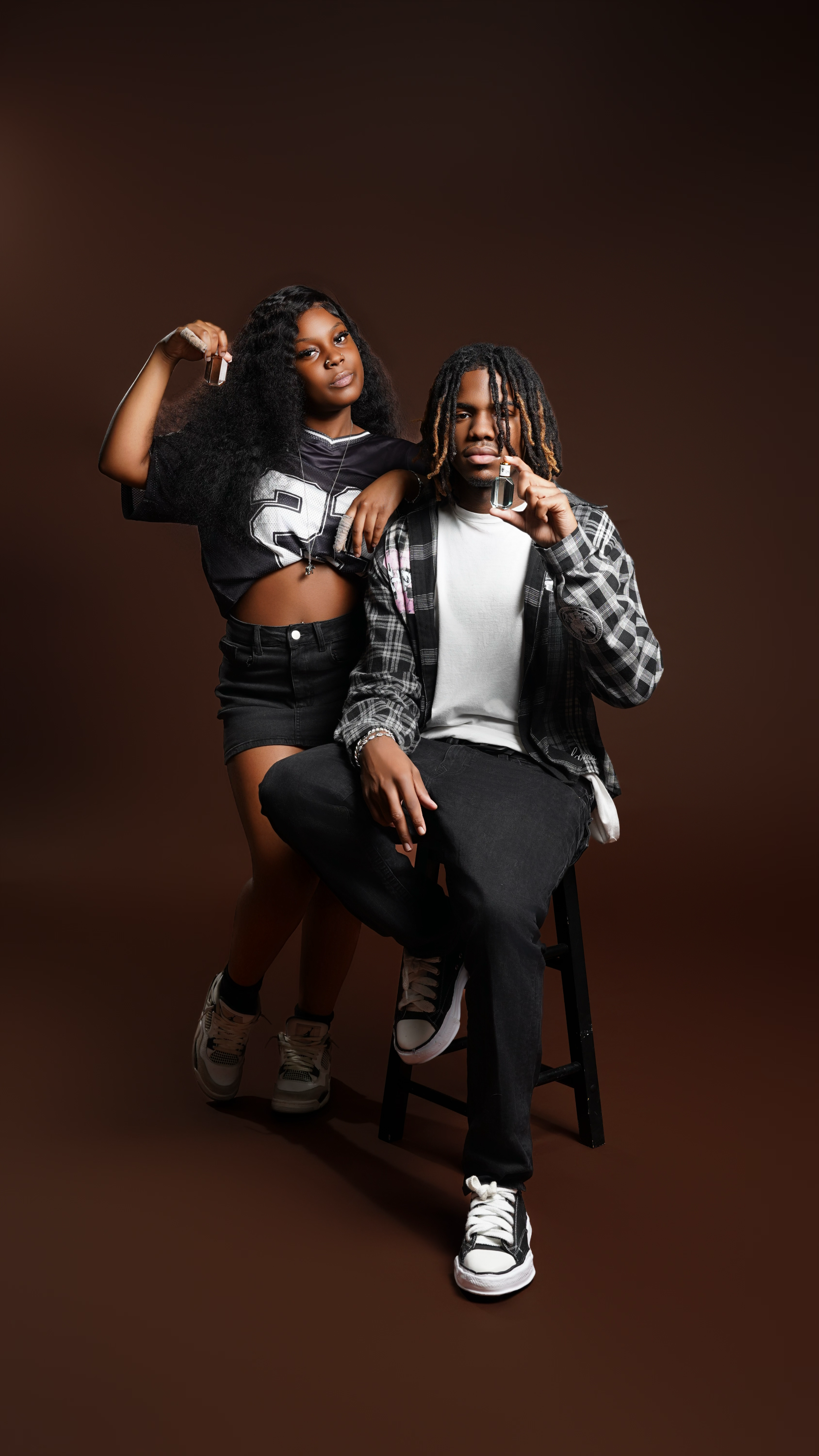 A young woman and man pose together with small liquor bottles in a studio against a brown background. The woman stands behind the man, resting her arm on his shoulder, while the man sits on a stool, holding a liquor bottle near his face.