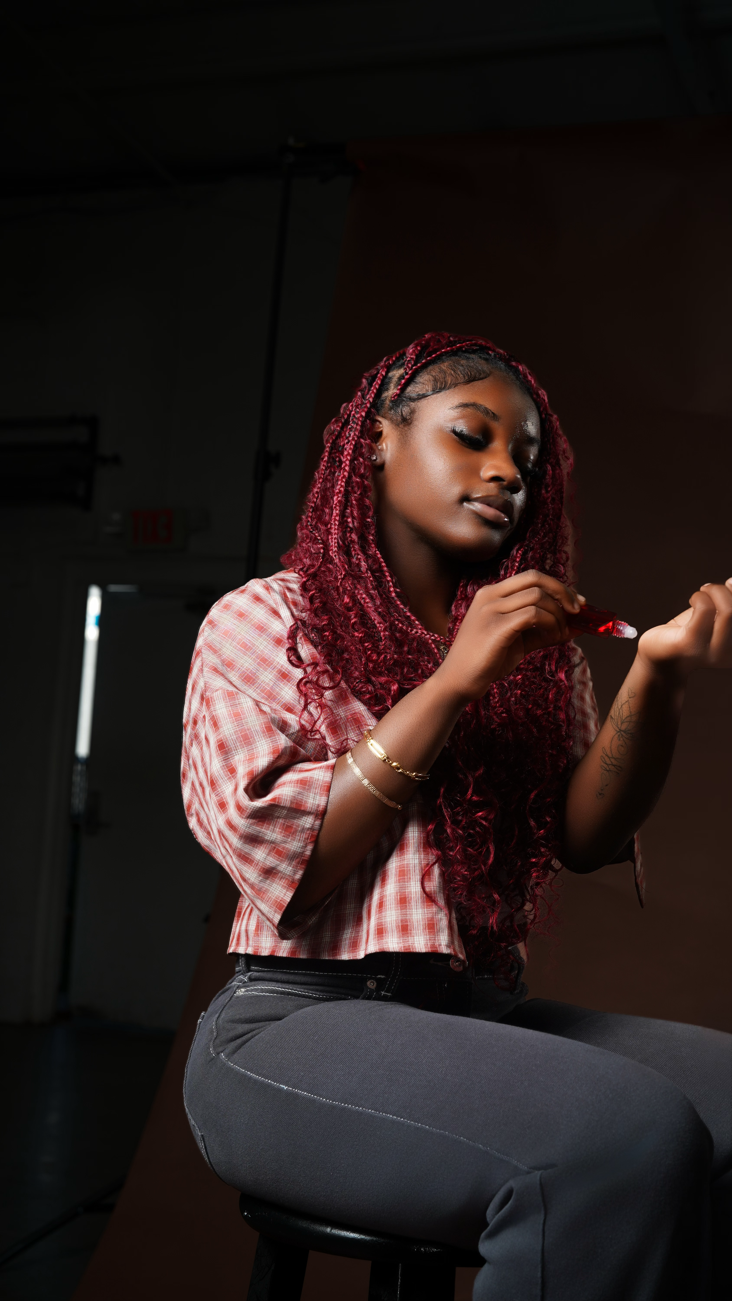 A young woman with long, curly, red-dyed hair applying lipstick in a photography studio.