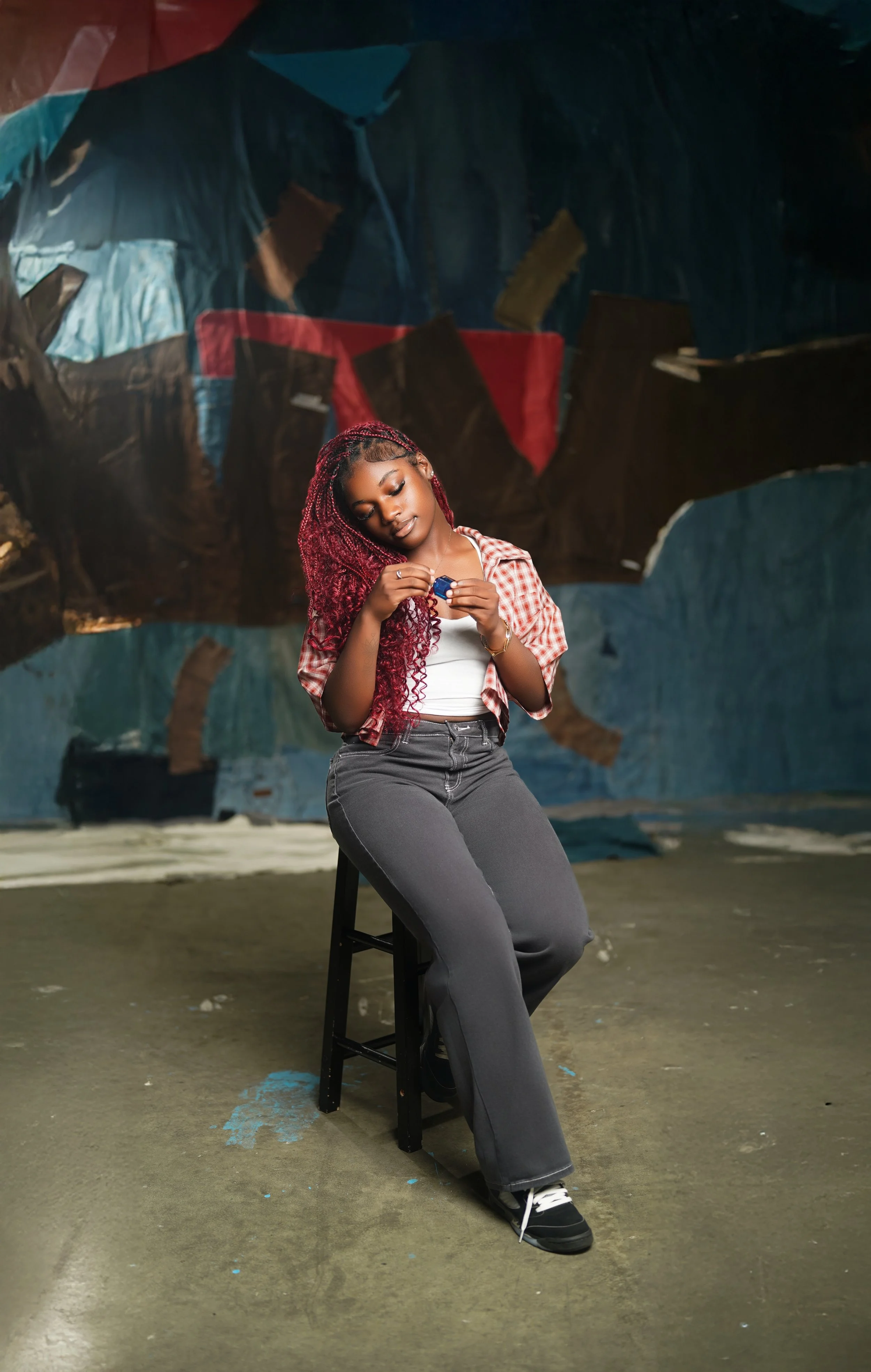 Young woman with red braided hair sitting on a black stool in front of a colorful abstract background, looking at a small object in her hands.