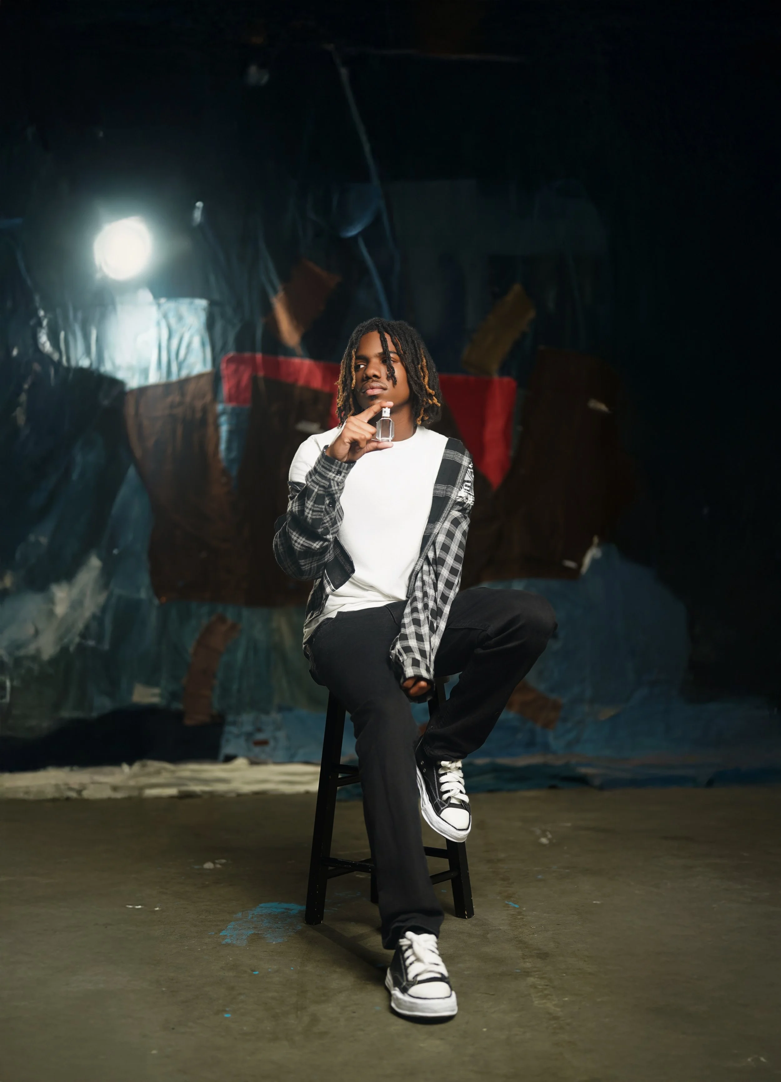 A young man with dreadlocks sitting on a black stool in an art studio with a large abstract painting in the background, holding a small bottle.