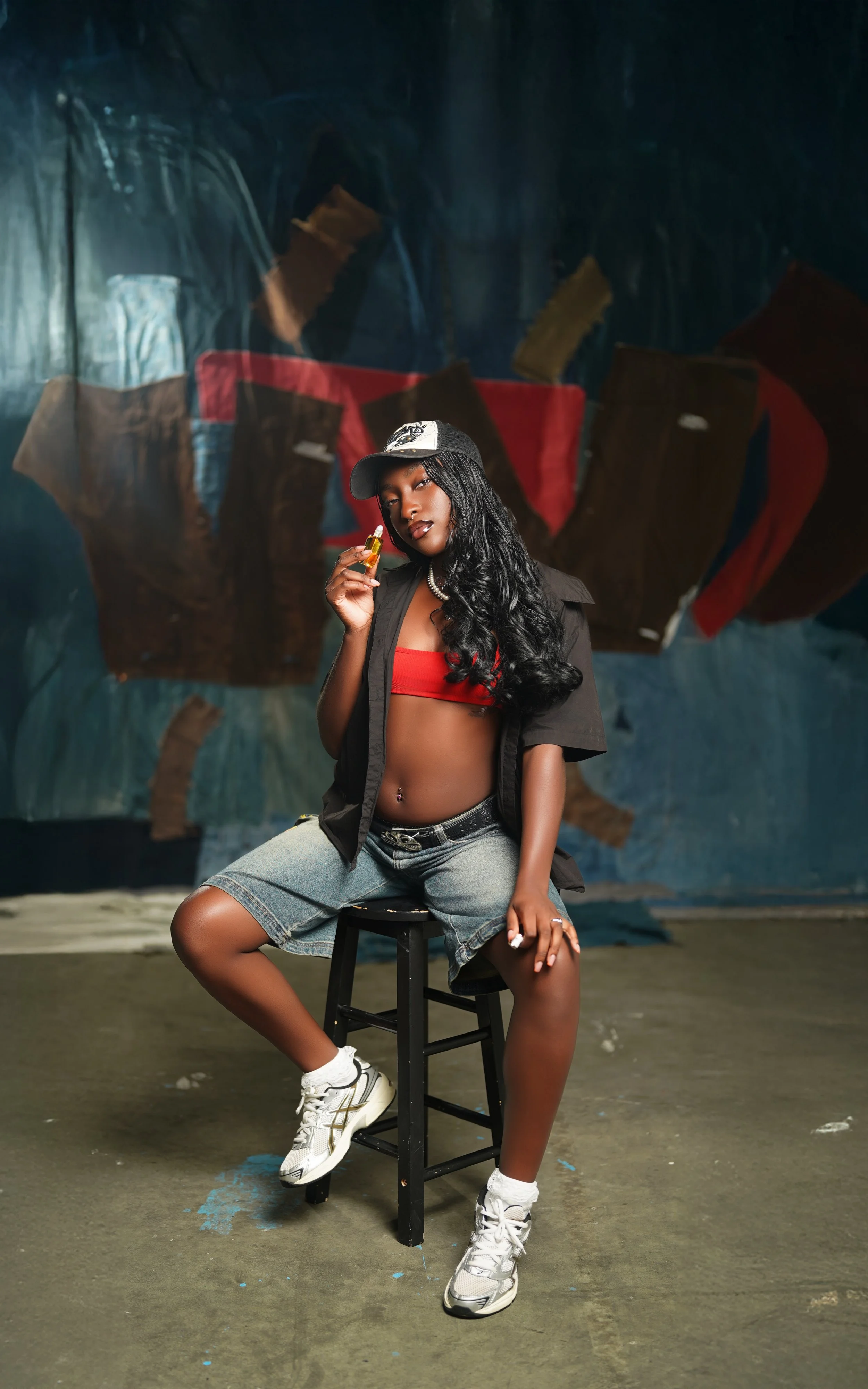 Young woman with long curly hair wearing a black cap, red crop top, black open shirt, denim shorts, and white sneakers, holding a vape pen, sitting on a black stool in front of an abstract, darkly painted background.