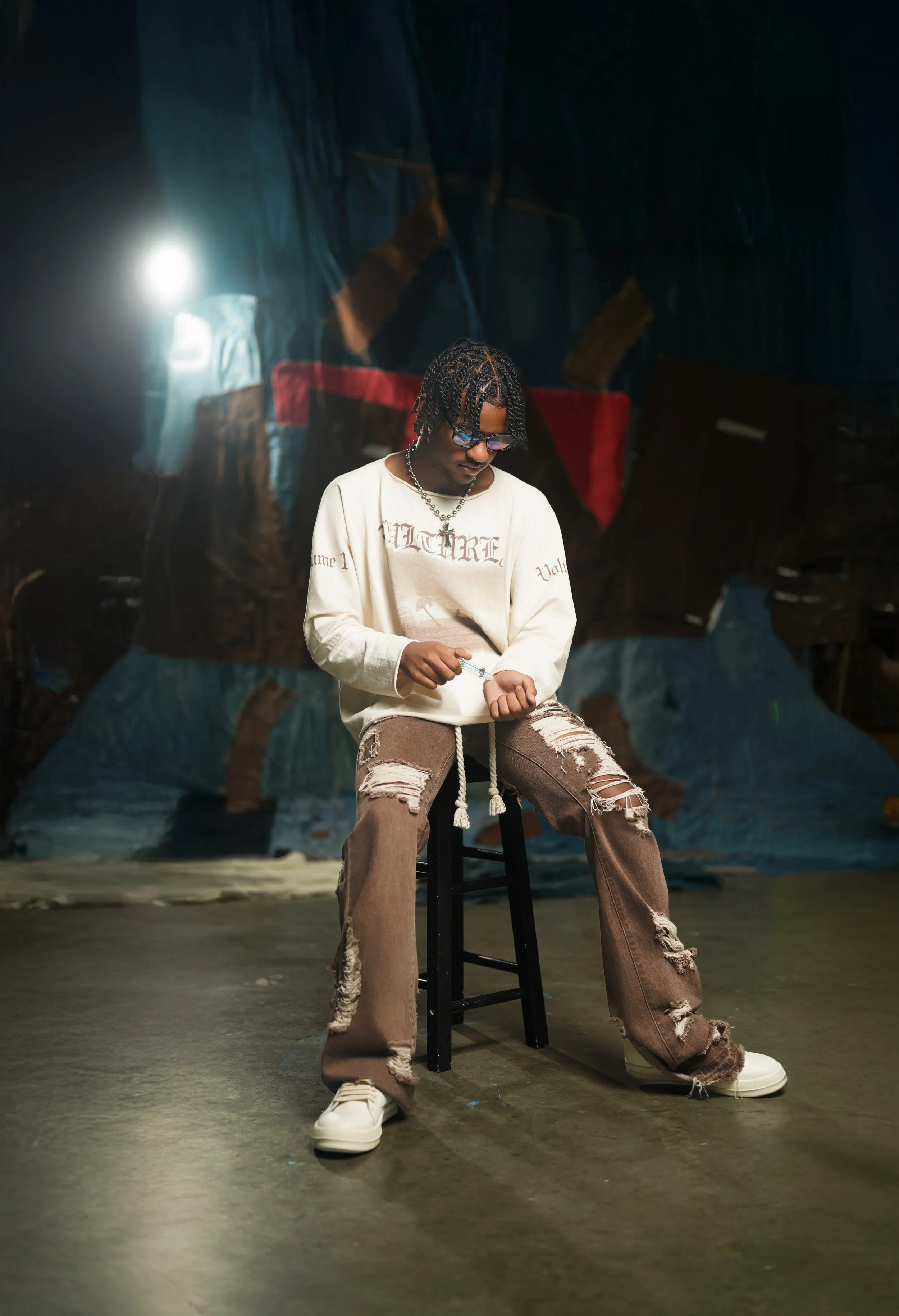 Young man with dreadlocks and glasses sitting on a black stool in a dimly lit artistic setting, wearing a beige oversized sweatshirt, ripped brown jeans, and white sneakers, holding a small object in his hands.