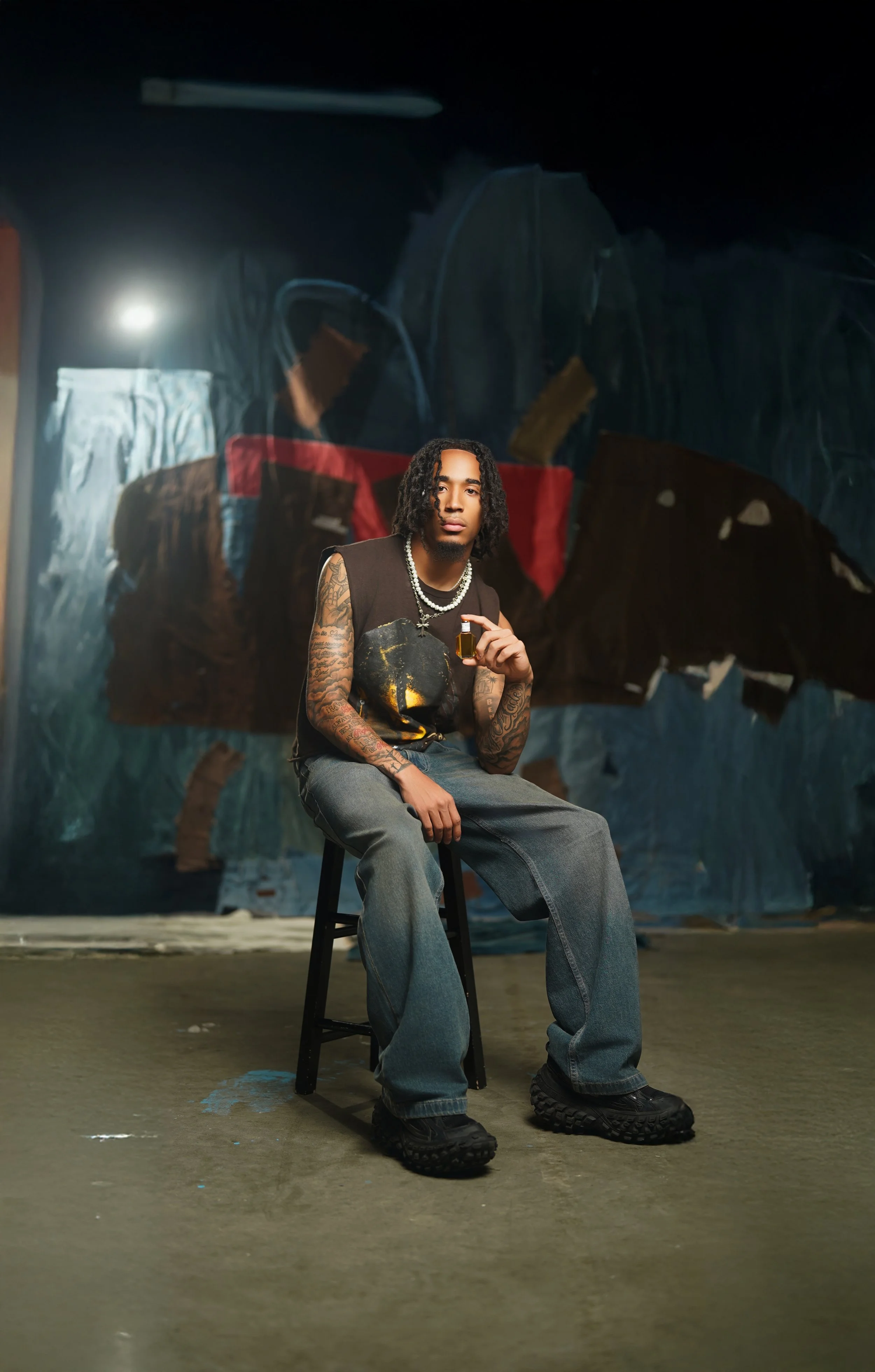 Young man with tattoos sitting on a black stool, holding a small bottle, in front of an abstract or painted backdrop.