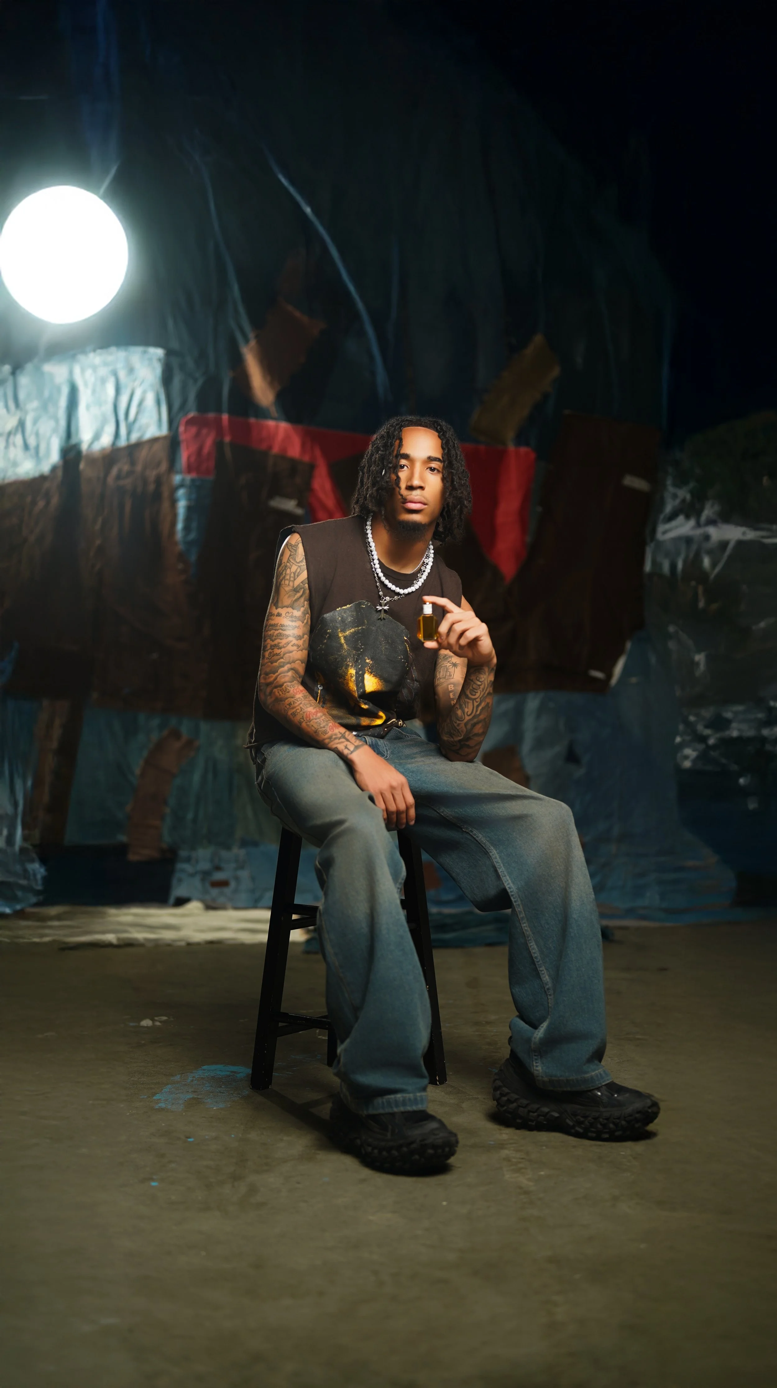A young man with tattoos, sitting on a black stool in a dimly lit room, holding a small bottle, with a dark, abstract background and a bright light to his left.