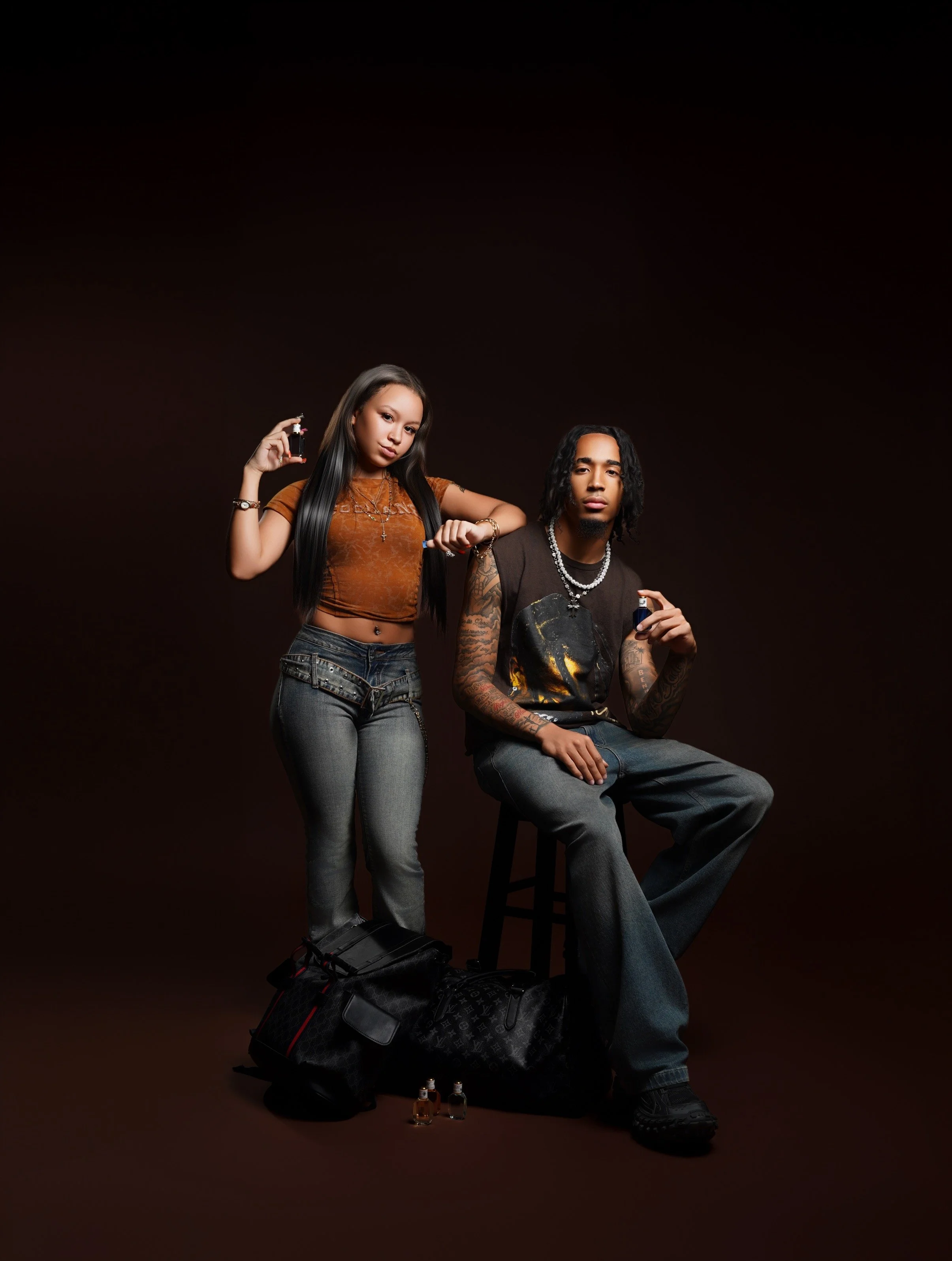 A young woman and man with dark hair and tattoos, posing against a black background. The woman is standing with one arm raised, holding a small bottle, and the man is sitting on a stool, also holding a small bottle. They are dressed in casual, edgy c