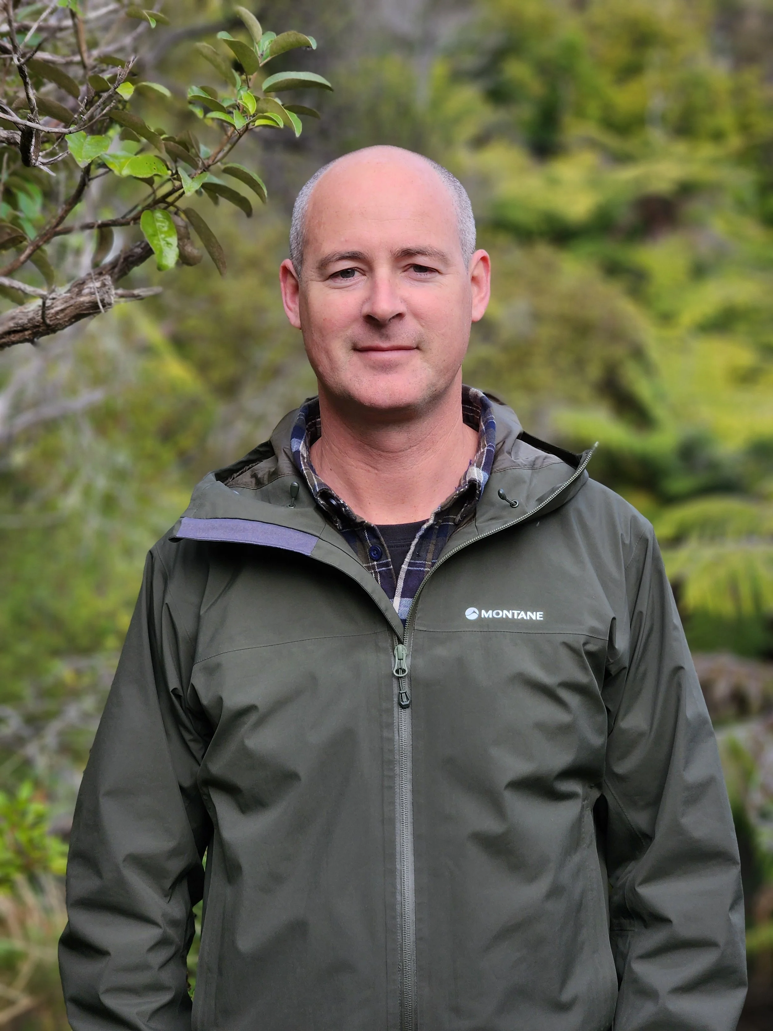 A man standing outdoors in a green natural setting, wearing a dark gray Montane jacket over a plaid shirt, with a calm expression.