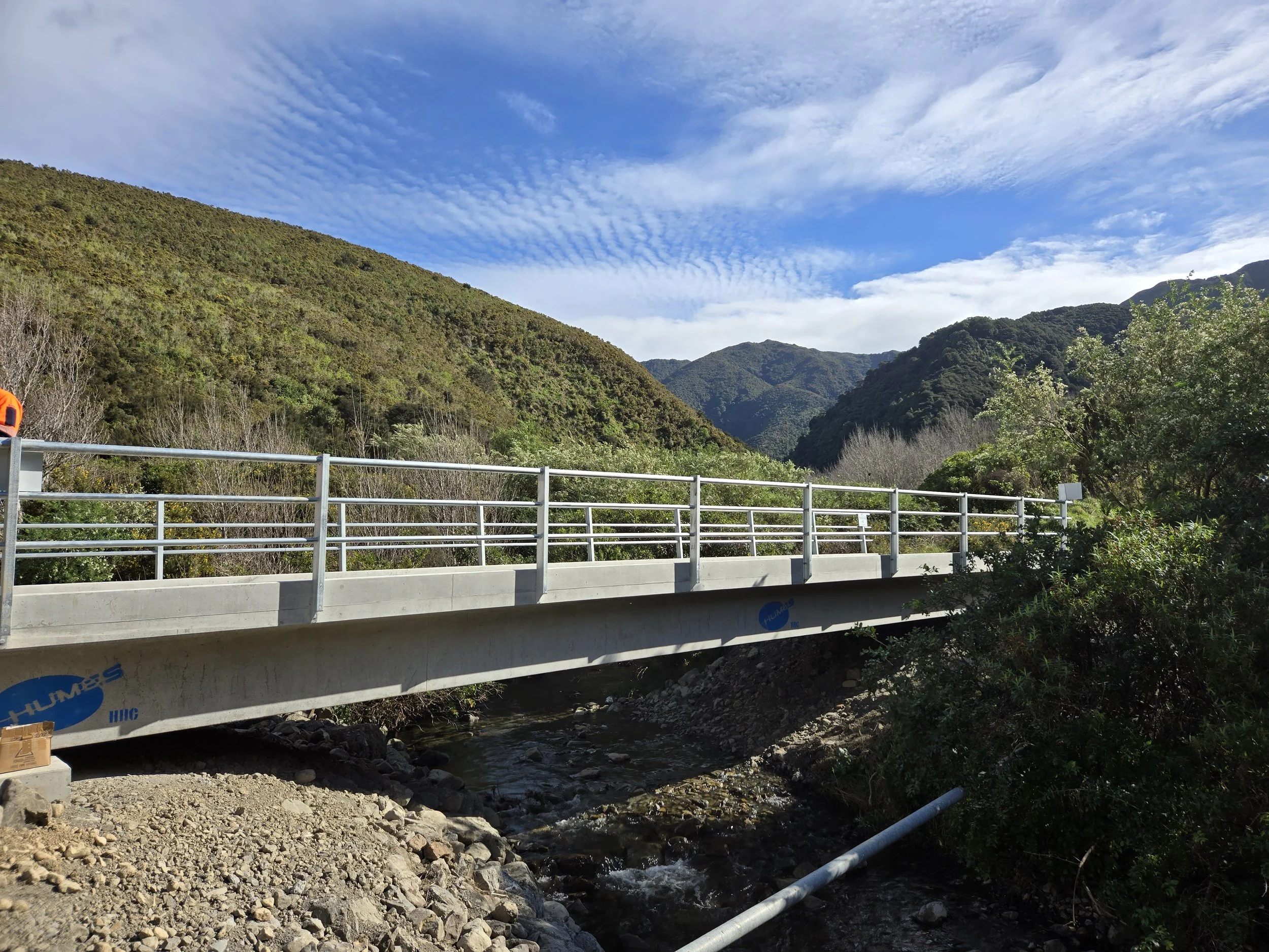 A bridge over a stream in a mountainous area with green hills, partly cloudy sky, and trees along the riverbank.